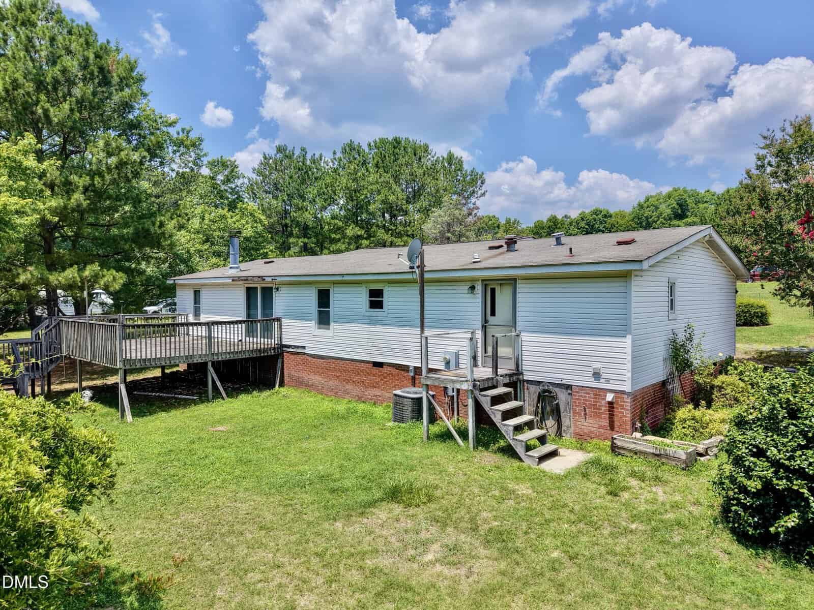 Single-story house with white siding and red brick foundation, wooden deck on the left, stairs leading to a grassy backyard, surrounded by trees, under a partly cloudy blue sky.