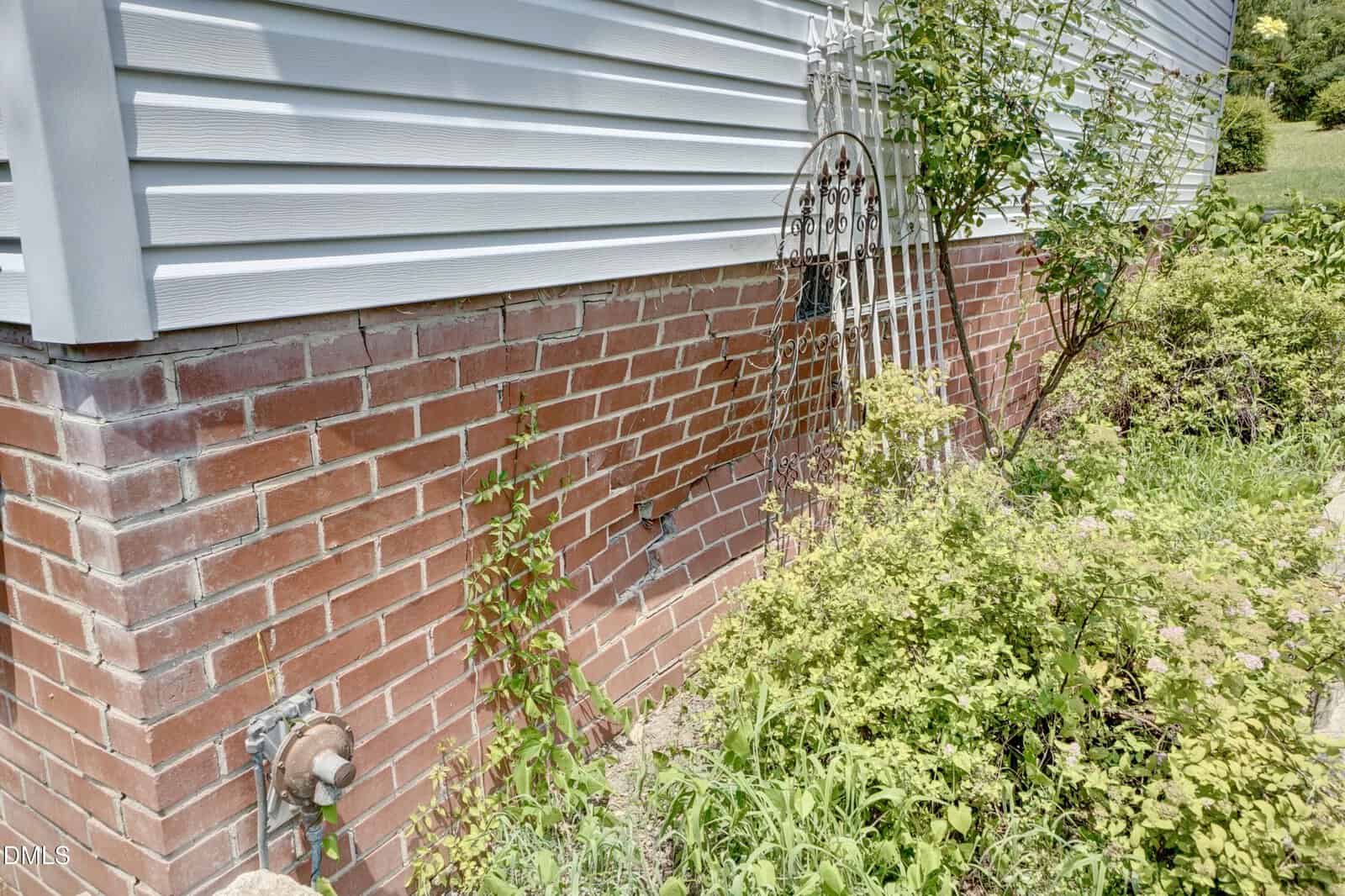 Side of a house with red brick foundation and white siding; overgrown plants and bushes line the base, with a metal trellis and a water spigot also visible.