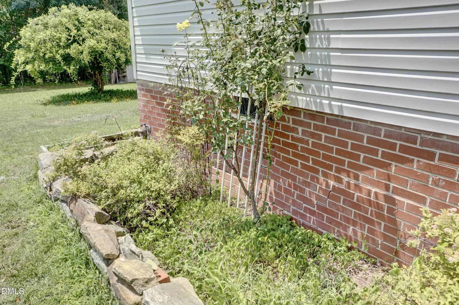 A brick house exterior with a small garden bed, green plants, a young rose bush, and rocks forming the garden border. Grass surrounds the garden on a sunny day.
