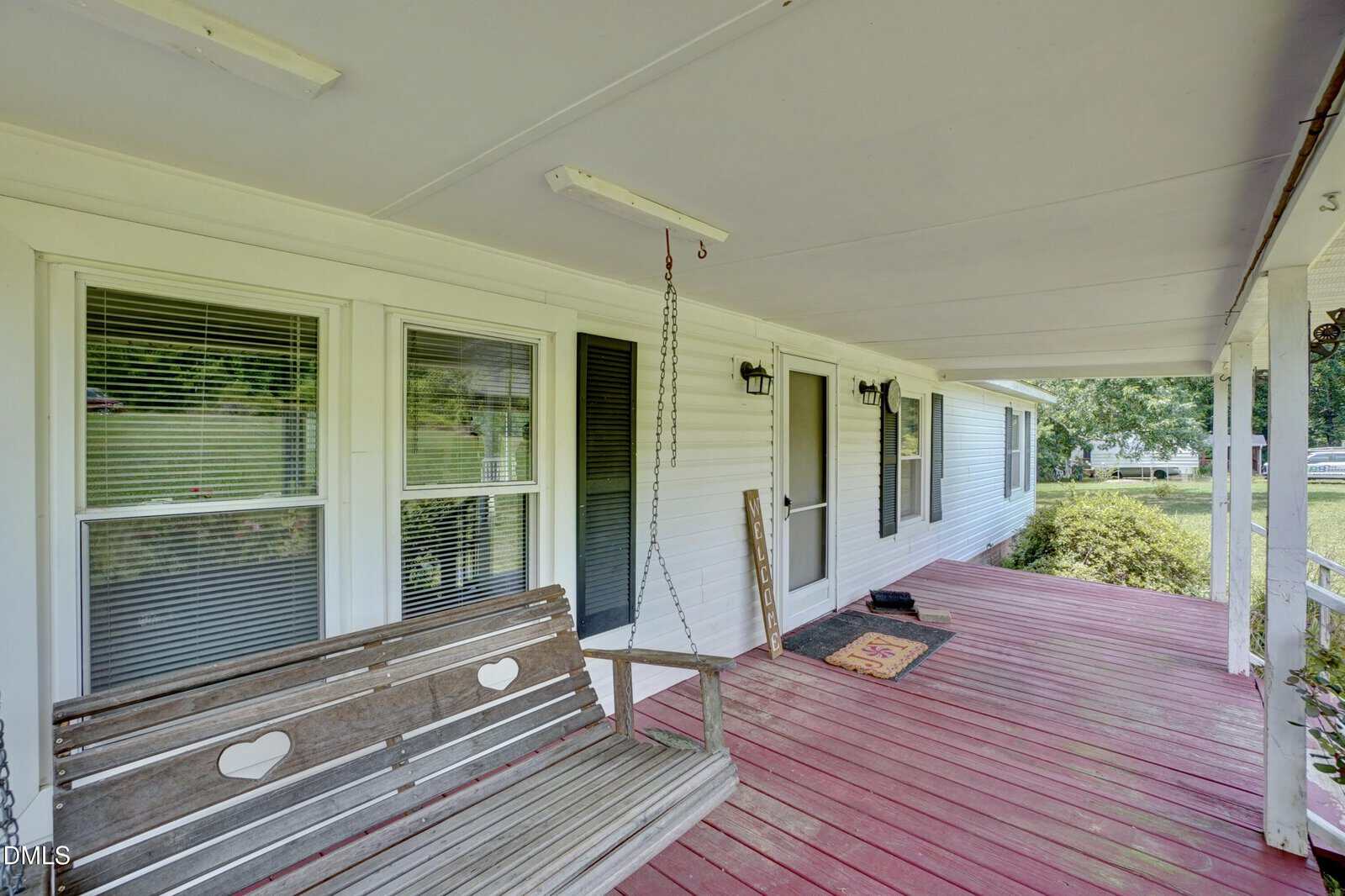 A wooden porch with red flooring features a hanging wooden swing, white railing, welcome mat, broom, and windows with blinds on a white house exterior. Green bushes and a lawn are visible.