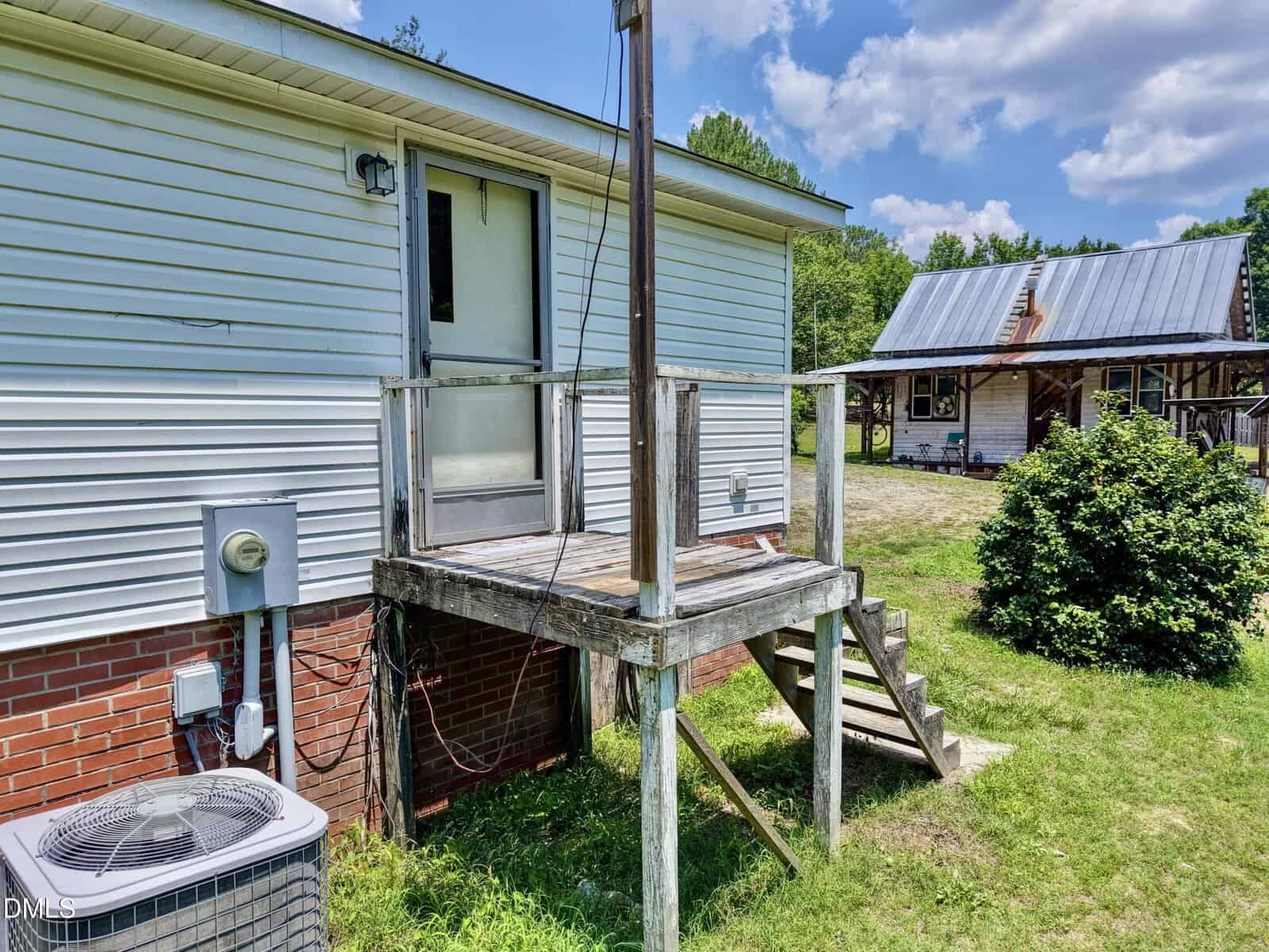 A small wooden porch with steps leads to a back door of a white house. There is an air conditioning unit below, a bush nearby, and another house with a metal roof in the background under a blue sky with clouds.