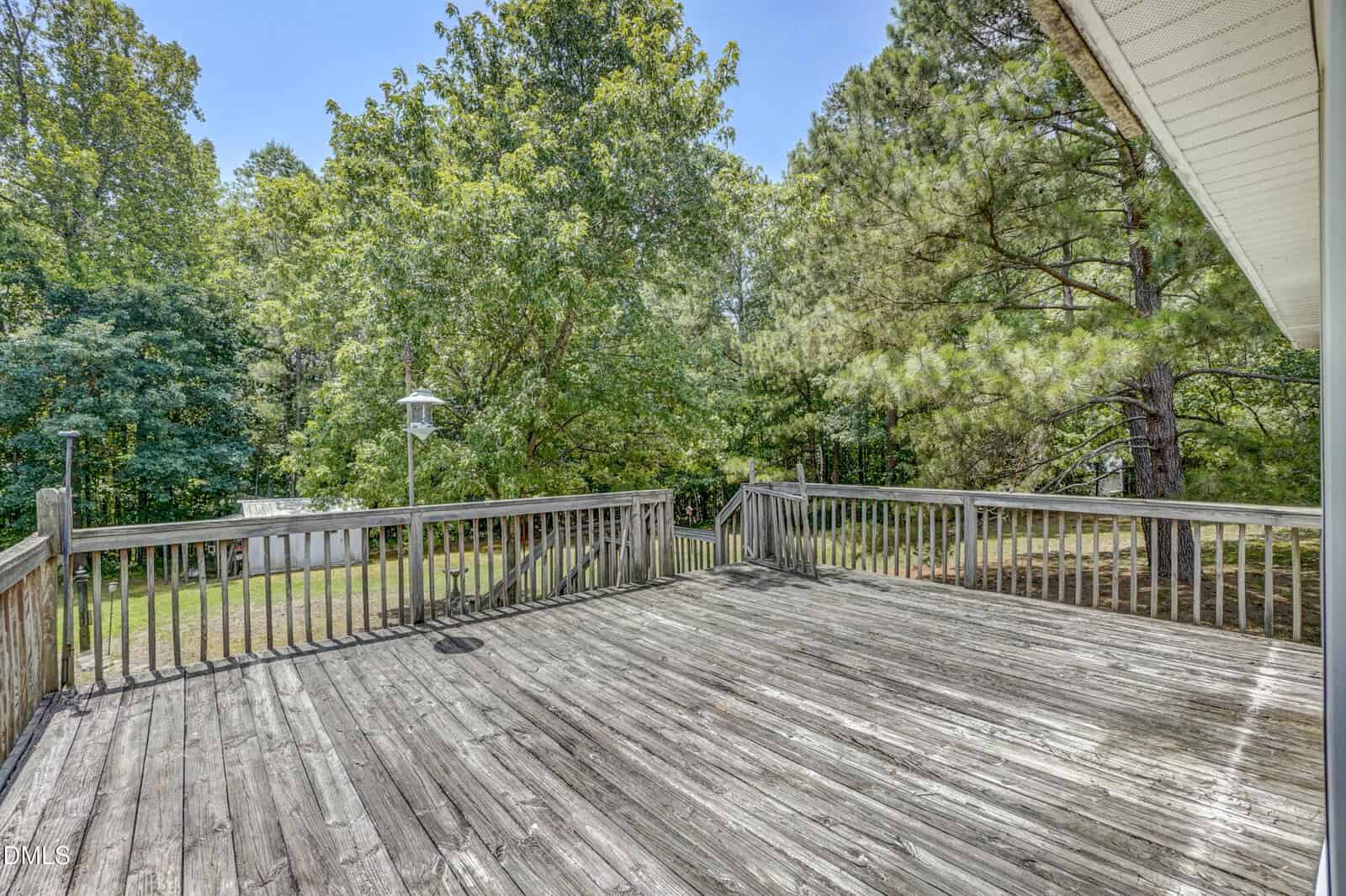 A spacious wooden deck with a weathered look overlooks a yard surrounded by tall green trees under a clear blue sky.