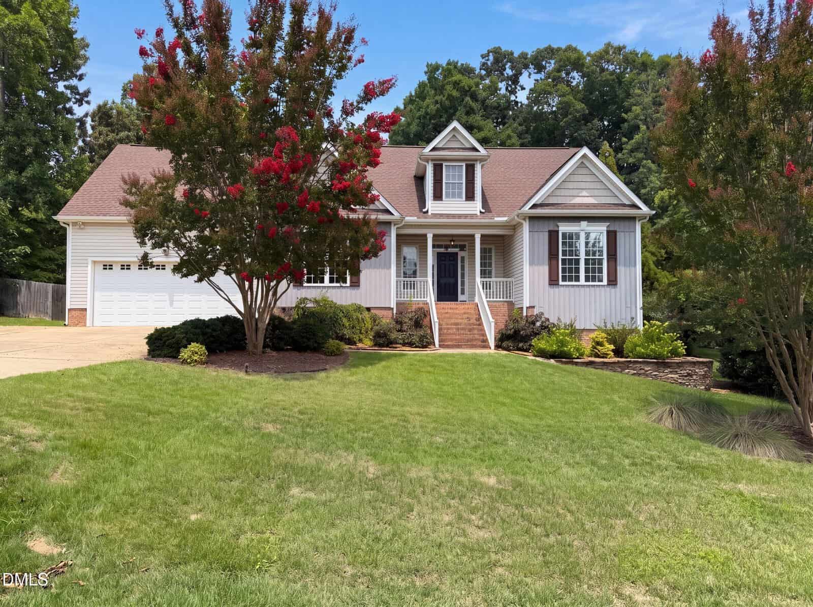 Charming single-story beige house with a gabled roof, a welcoming front porch, and two blooming crepe myrtle trees gracing the green lawn. This inviting residence at 40 Red Rock Ridge in Youngsville also features an attached two-car garage and is set against a picturesque backdrop of tall trees and blue sky.