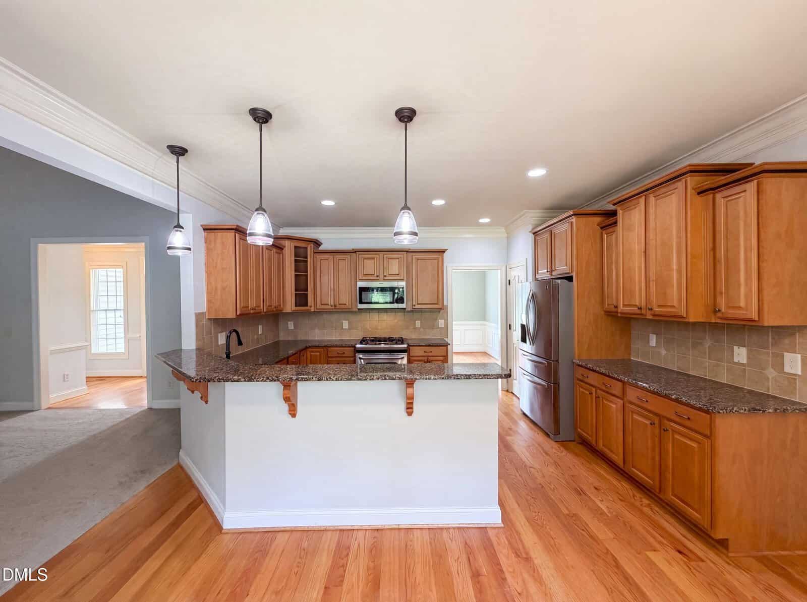 Spacious kitchen with wooden cabinets, granite countertops, stainless steel appliances, a breakfast bar, pendant lighting, and hardwood flooring. An adjacent room and hallway are visible in the background.