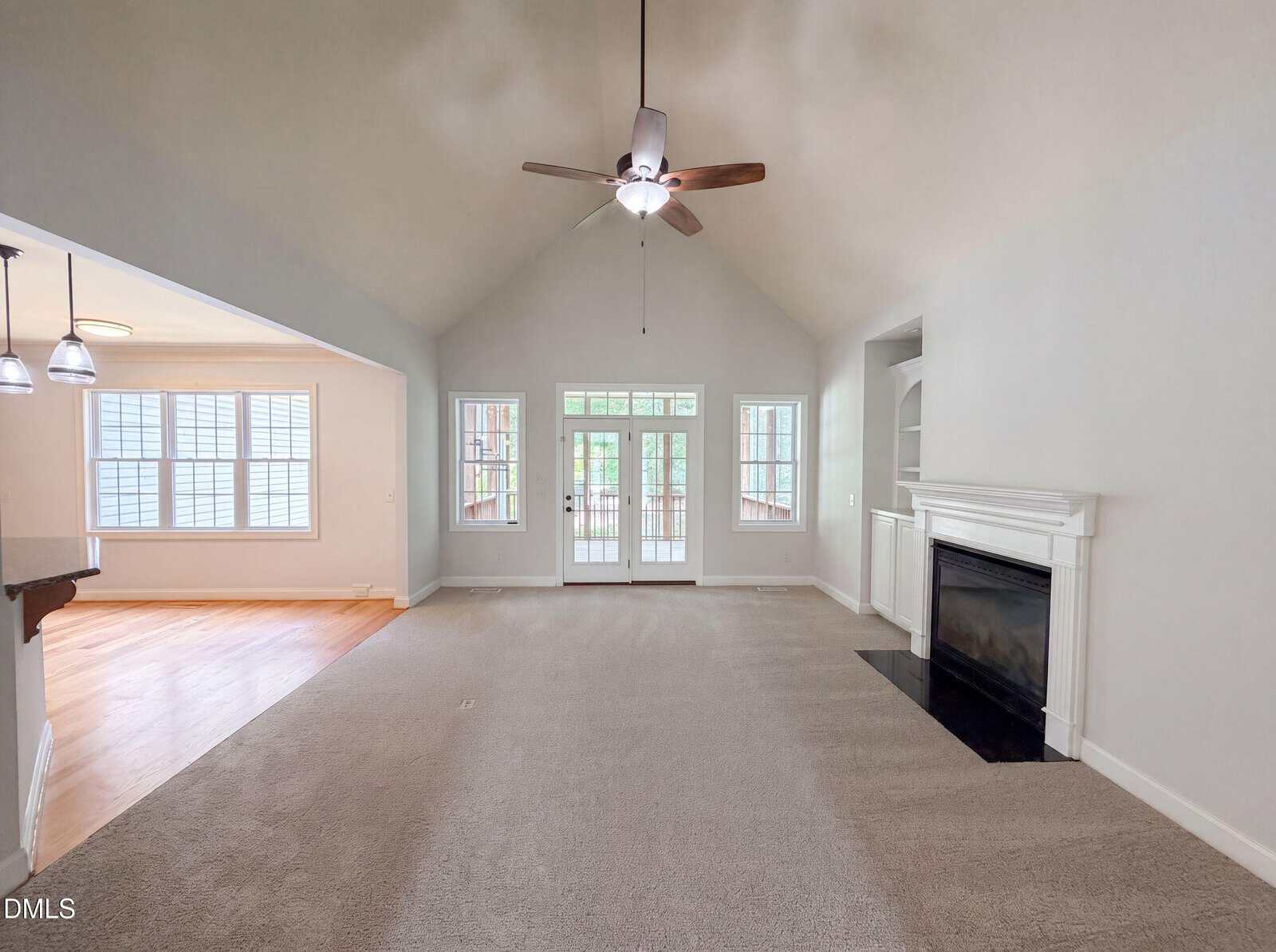 Spacious living room with vaulted ceiling, ceiling fan, carpet, fireplace, large windows, and glass doors leading to a sunroom. Adjoining area has hardwood floors and pendant lights over a breakfast bar.
