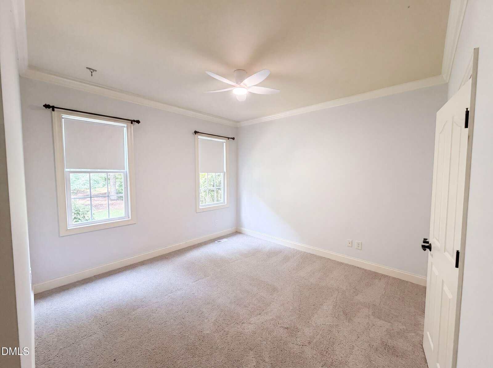 Bright, empty bedroom with two windows, beige carpet, light-colored walls, white ceiling fan, and a closed white door. Natural light enters through the windows, and outdoor greenery is visible.