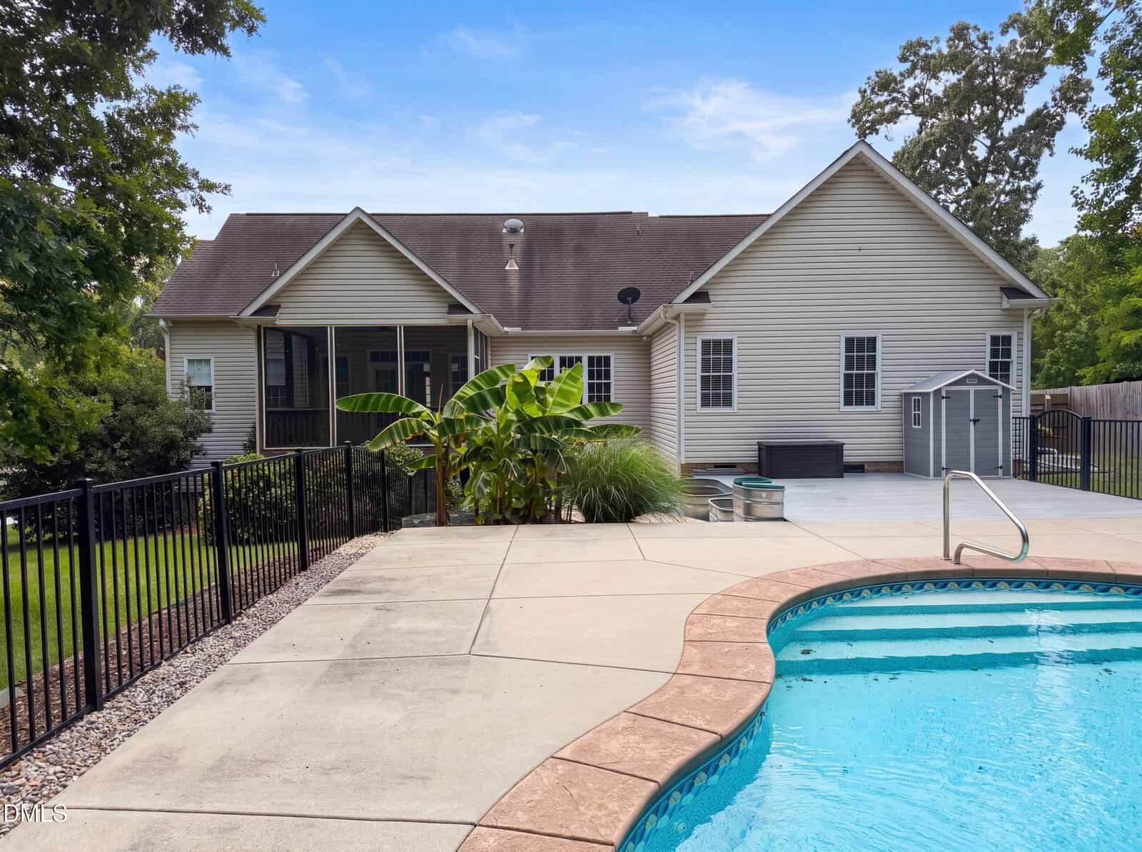 A backyard with a swimming pool, concrete patio, tropical plants, a small shed, and a beige house with multiple windows under a blue sky.