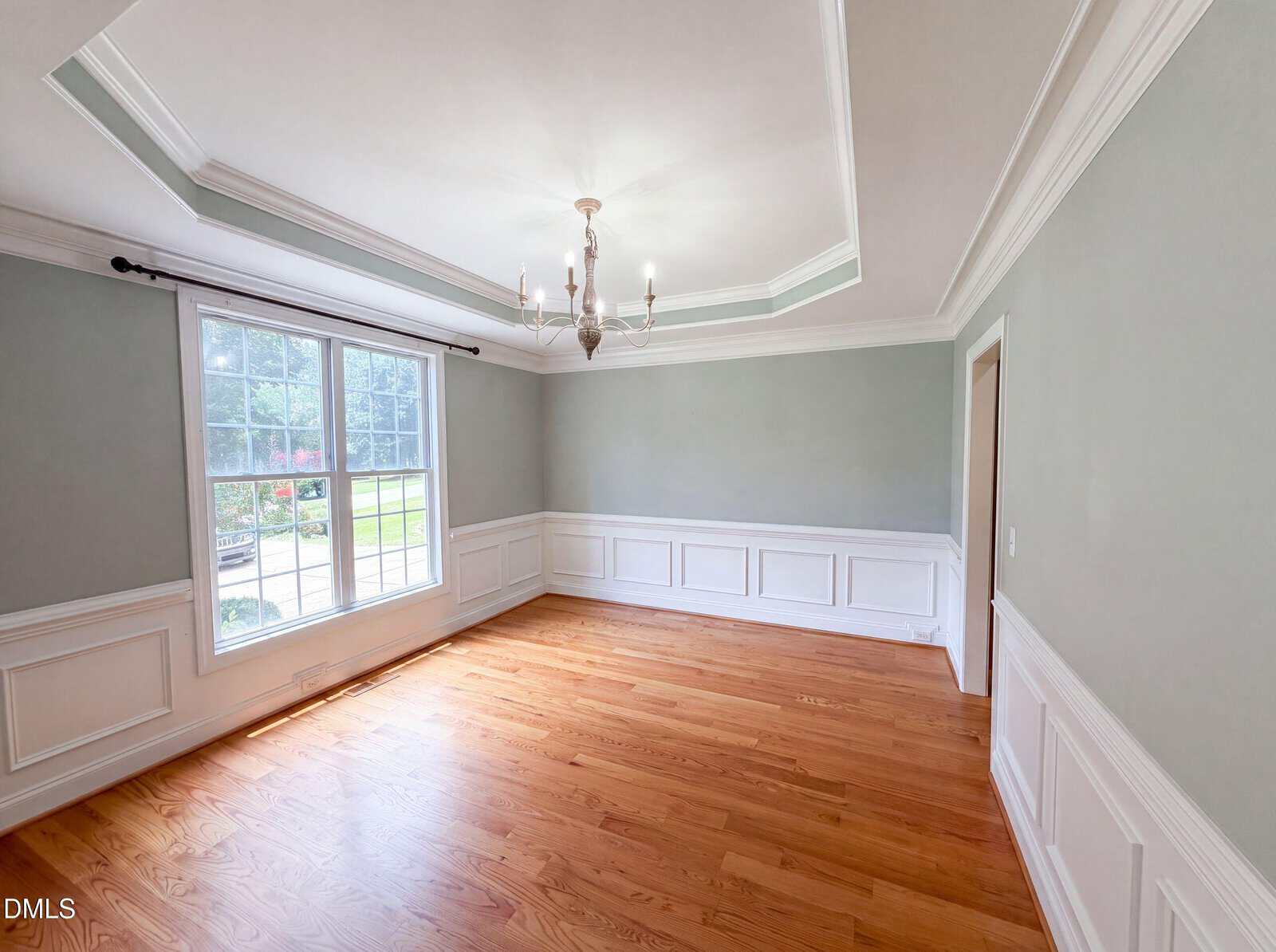 Empty room with light green walls, white wainscoting, hardwood floor, large window with outside view, and a ceiling chandelier. Natural light fills the space.