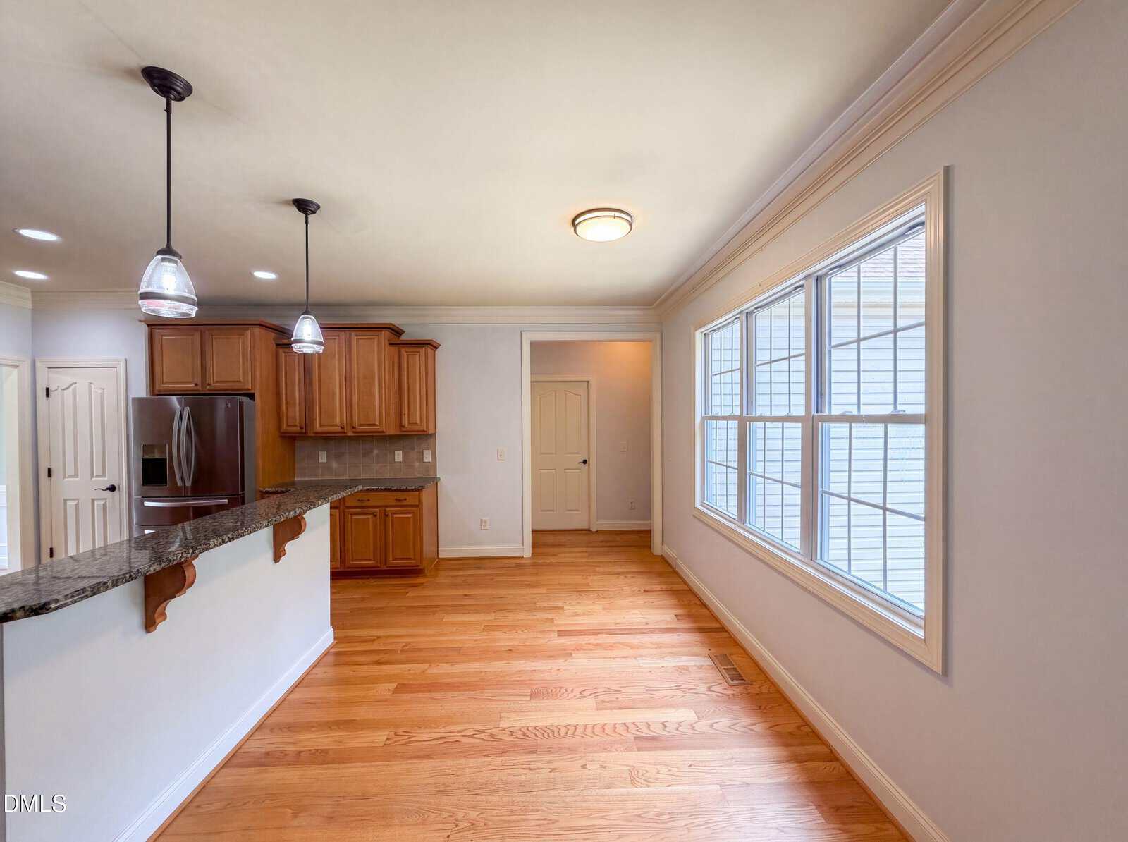 Kitchen with wooden cabinets, stainless steel appliances, granite countertop with a bar area, pendant lights, hardwood floors, large windows on the right, and a white door at the back. Walls are light-colored.