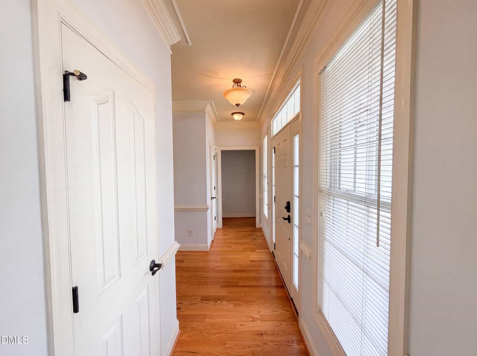 A hallway with light wood floors, white walls, three doors on the left, and two doors with glass panels and blinds on the right; a ceiling light fixture is visible at the end.