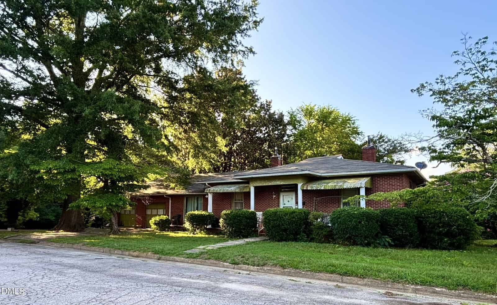 A single-story red brick house with a flat roof, surrounded by green bushes and trees, sits on a quiet street with a cracked, paved surface under a clear blue sky.