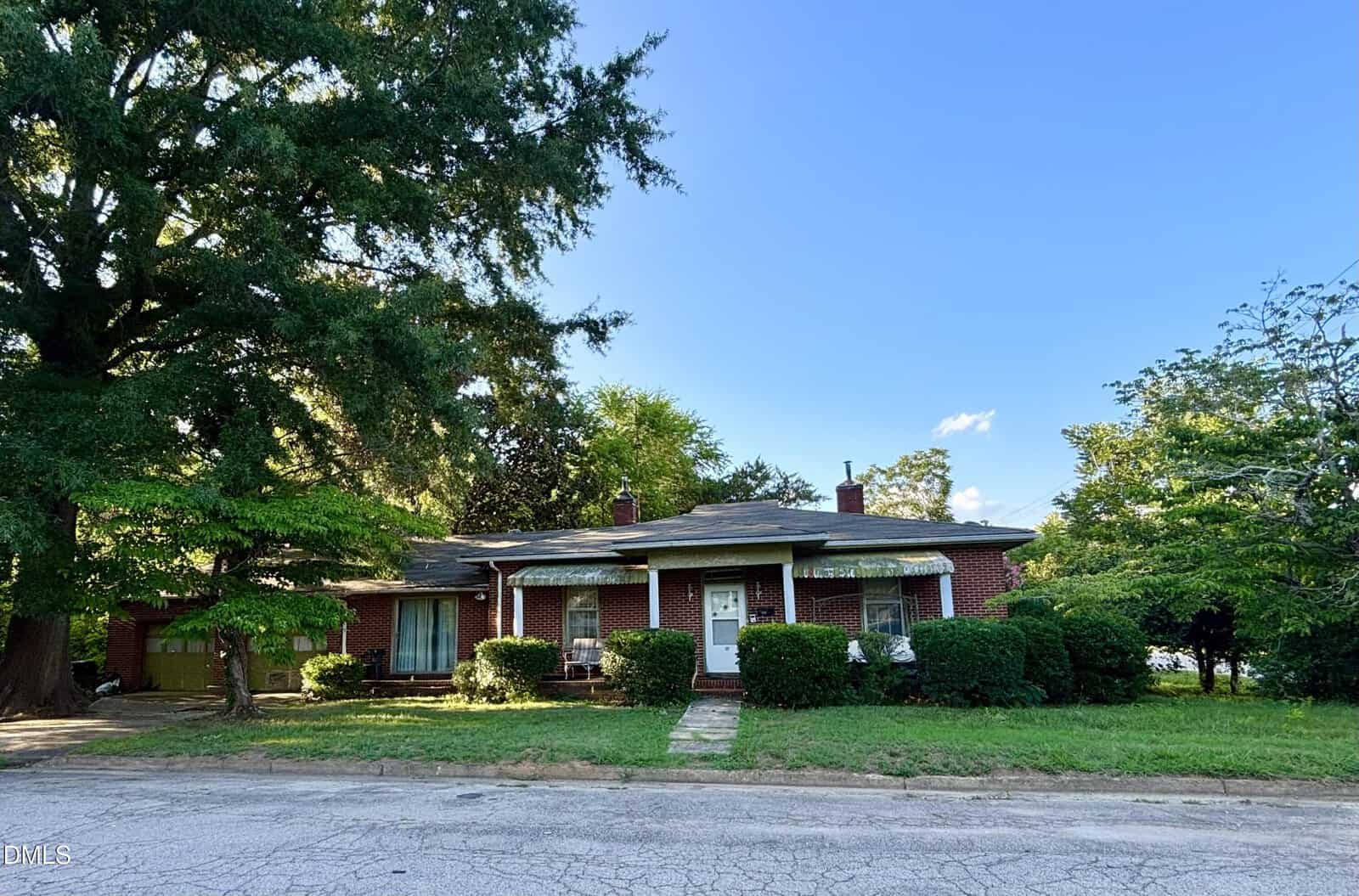 A one-story red brick house with white trim, two front doors, small porches, and bushes out front sits beneath a large leafy tree on a quiet street in Warrenton at 114 College Street, all under a clear blue sky.