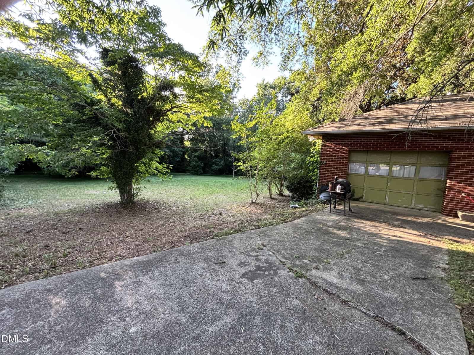 A concrete patio leads to a grassy backyard surrounded by trees. A red brick garage with green doors sits on the right, and a small grill is visible near the garage. Sunlight filters through the trees.