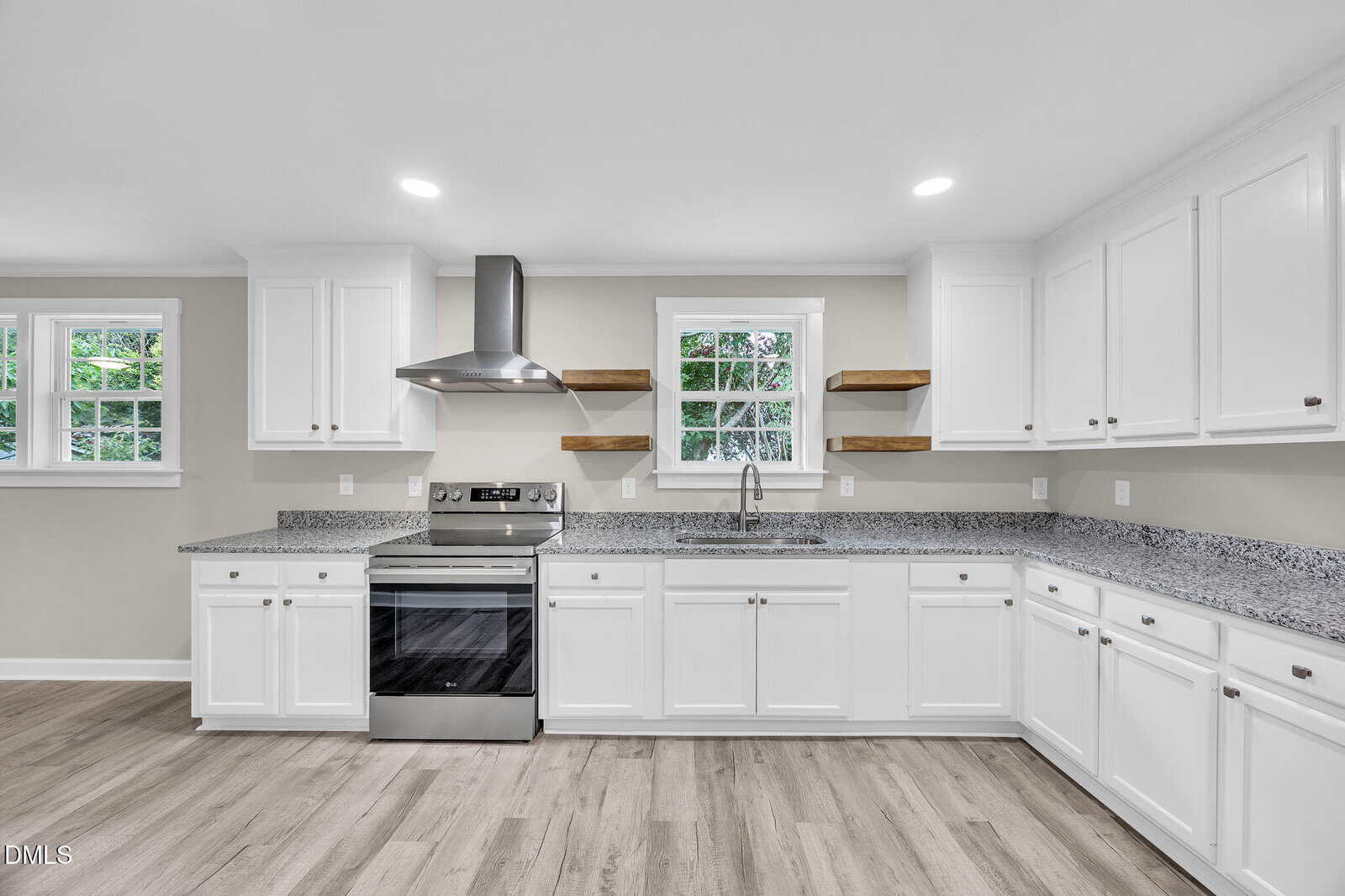 Modern kitchen with white cabinets, stainless steel stove and hood, granite countertops, two wooden open shelves, two windows, and light wood flooring. The space is bright and uncluttered.