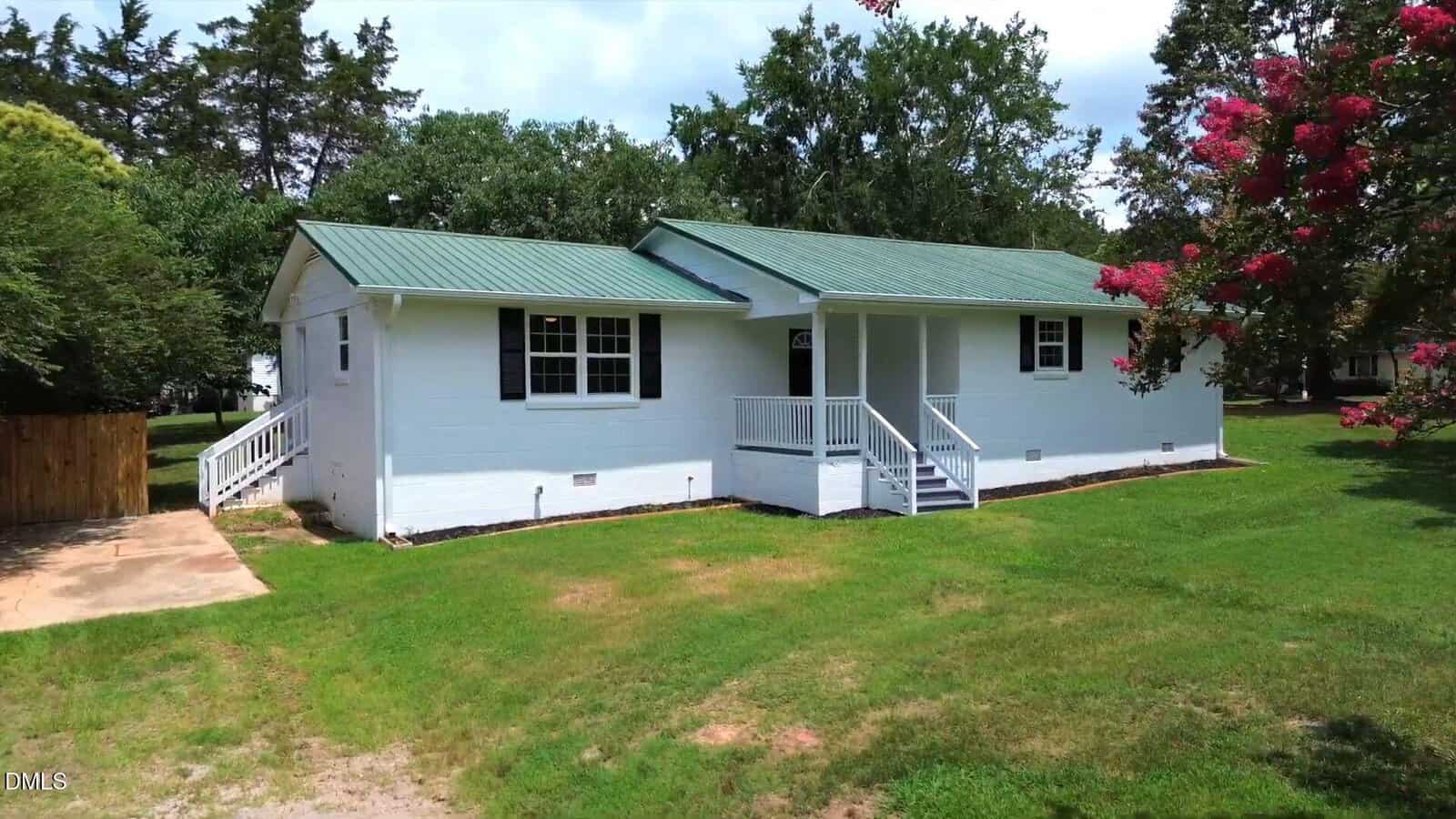 Charming white single-story house at 501 Warren Street in Warrenton, featuring a green metal roof, black shutters, and a cozy front porch. Surrounded by lush green grass and mature trees, the home offers inviting outdoor spaces and a convenient driveway on the left side.