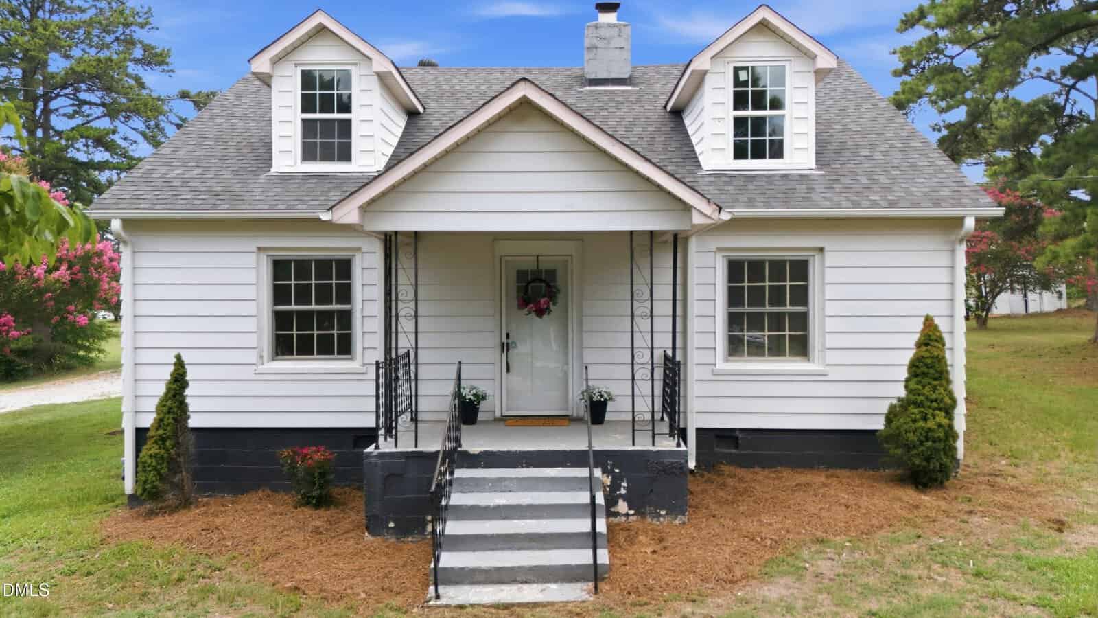 White house with two dormer windows, a covered front porch, black railings, and steps leading to a white front door with a wreath. Two small shrubs and flower pots are near the entrance. Green lawn and trees surround the house, offering inviting curb appeal at 5083 NC 56 Hwy in Franklinton.