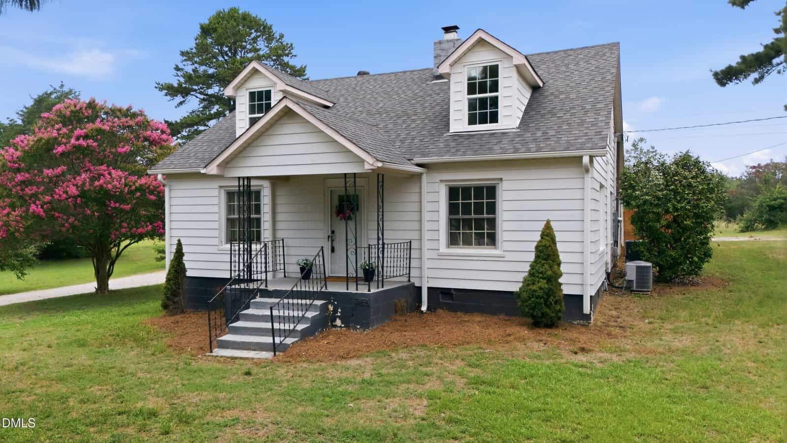 A white, two-story house with gray roof, covered front porch, black railing, and two dormer windows. The house is surrounded by grass, trees, and flowering bushes under a blue sky.