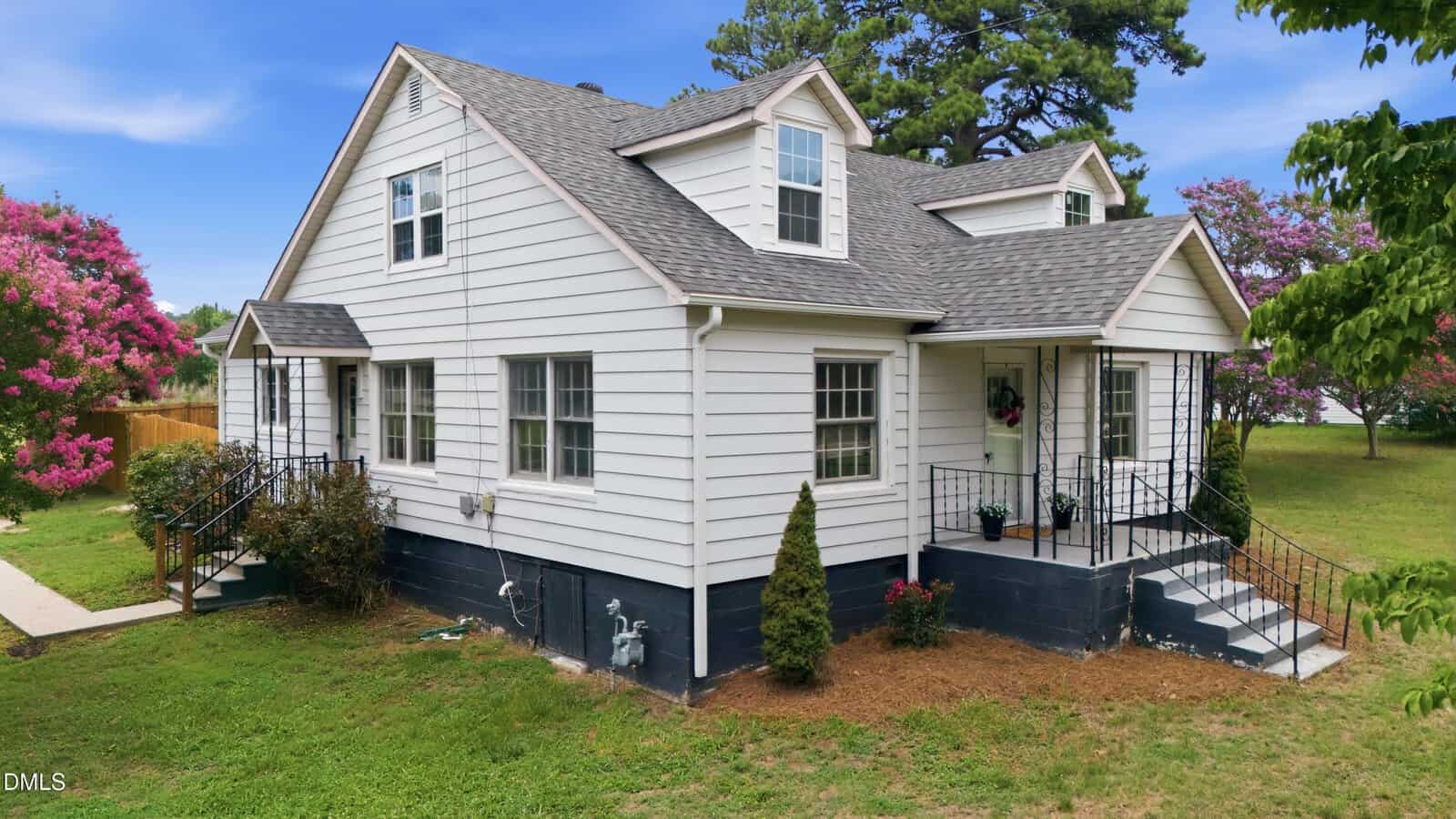 White two-story house with a gray roof, black foundation, small front porch, and metal railings. Surrounded by green grass, bushes, and pink flowering trees. A sidewalk leads to a side entrance.