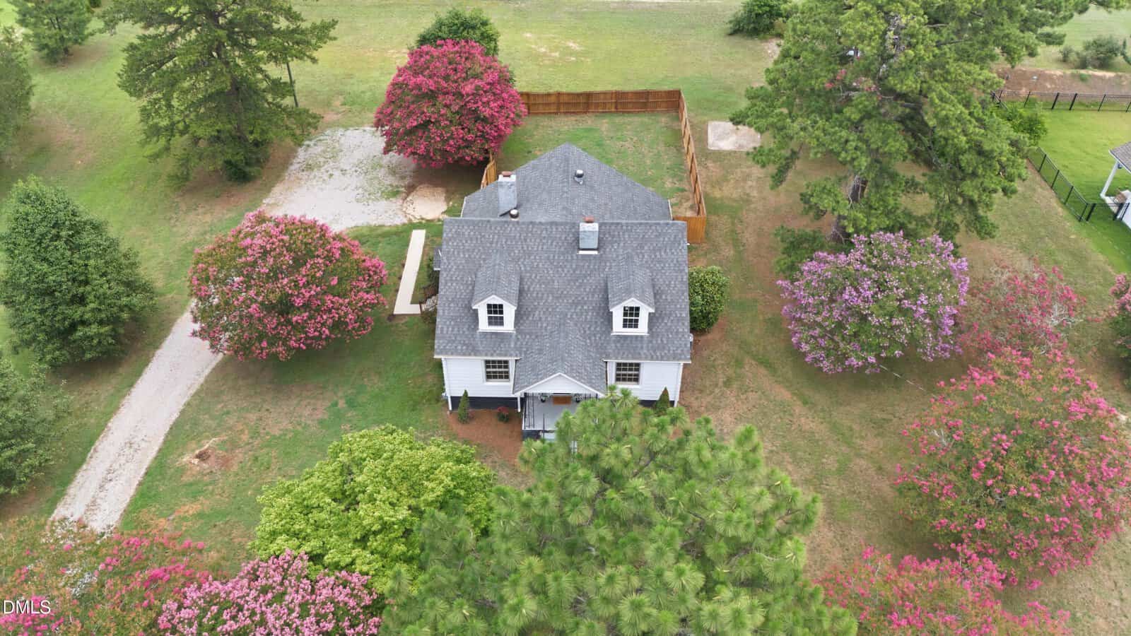 Aerial view of a white house with a gray roof, surrounded by blooming pink and purple trees, a gravel driveway, green lawn, and fenced backyard.