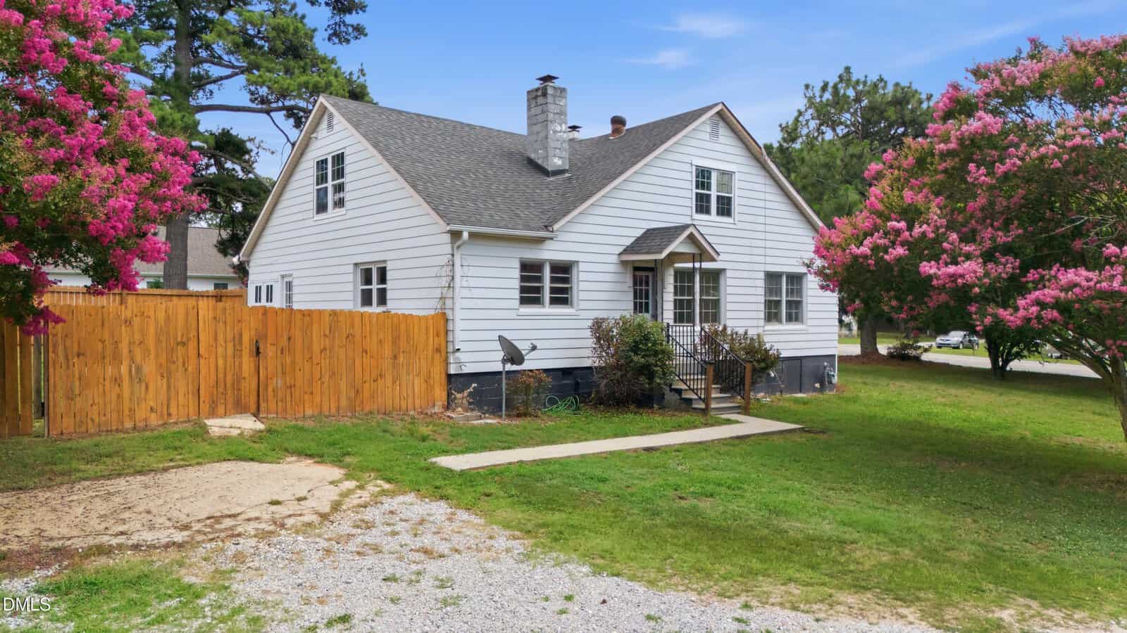 A white, two-story house with a gray roof and chimney sits on a grassy lot, bordered by pink flowering trees and a wooden fence, under a blue sky.