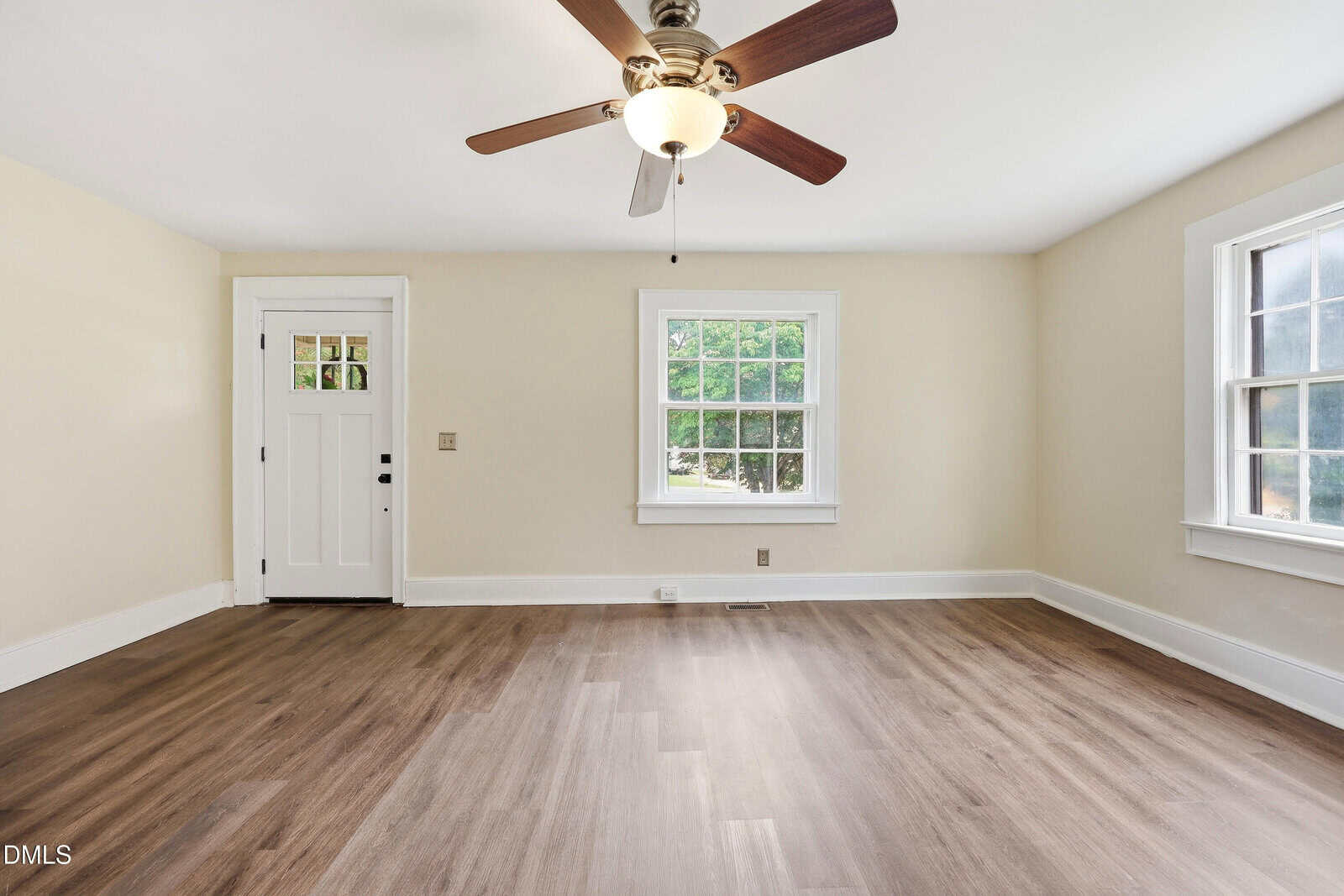 A bright, empty room with light beige walls, a ceiling fan, wood-look flooring, and two windows letting in natural light. There is also a white door on the left wall.