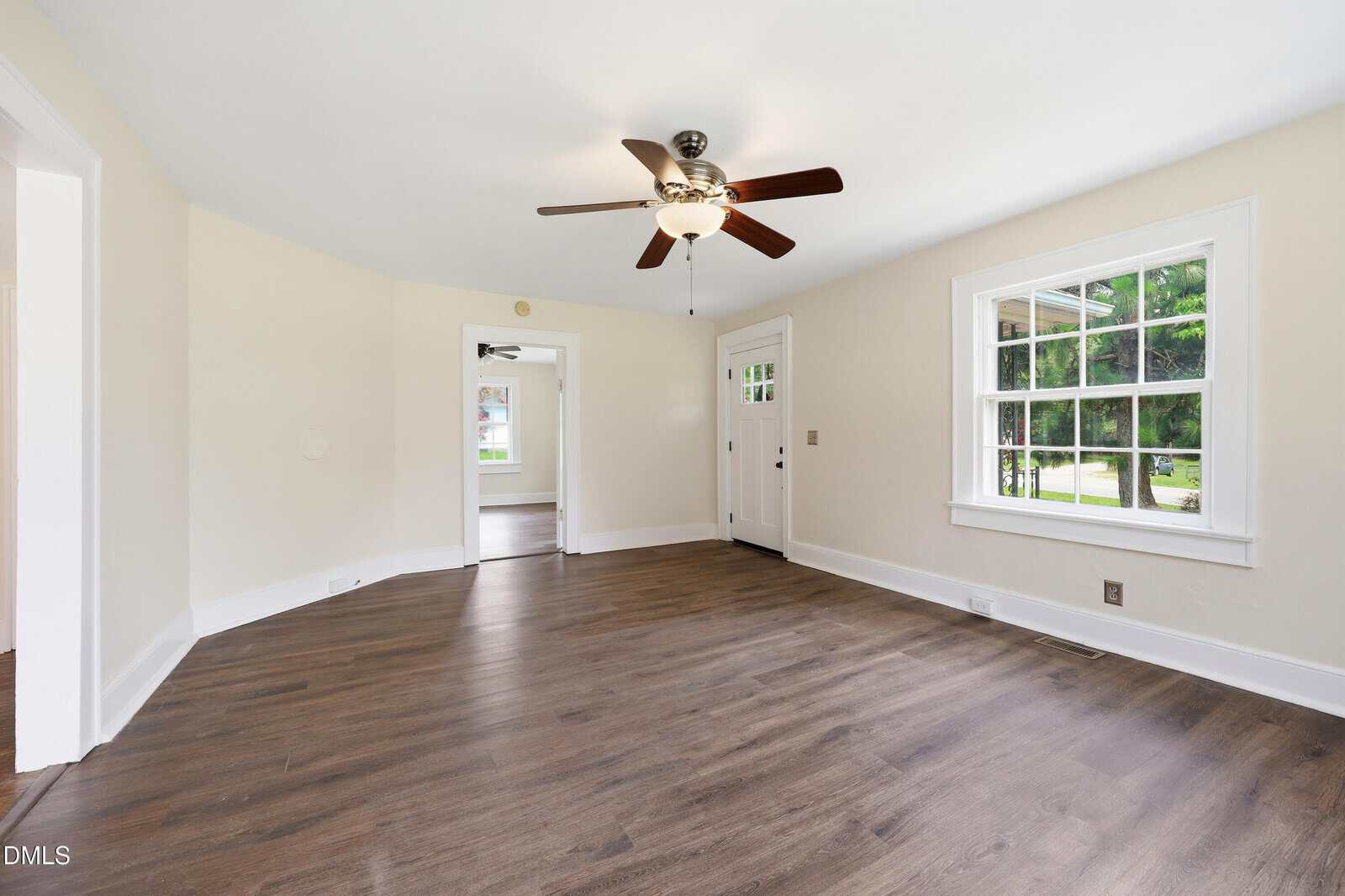 Bright, empty living room with wood floors, white walls, large front window, ceiling fan with lights, and an open doorway leading to another room.