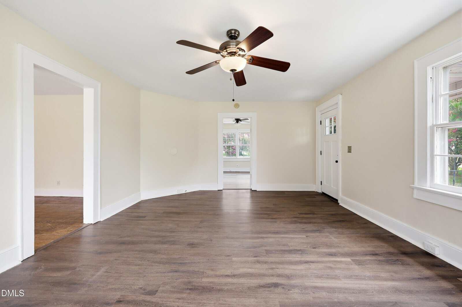 Empty living room with hardwood floors, beige walls, white trim, a ceiling fan, a door with windows, and large windows letting in natural light. Two doorways lead to adjacent rooms.