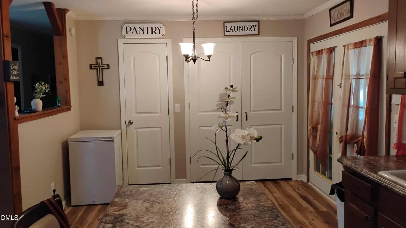 A cozy kitchen with double doors labeled "PANTRY" and "LAUNDRY," a small freezer, a cross on the wall, brown curtains, and a dining table with a vase of white orchids in the center.