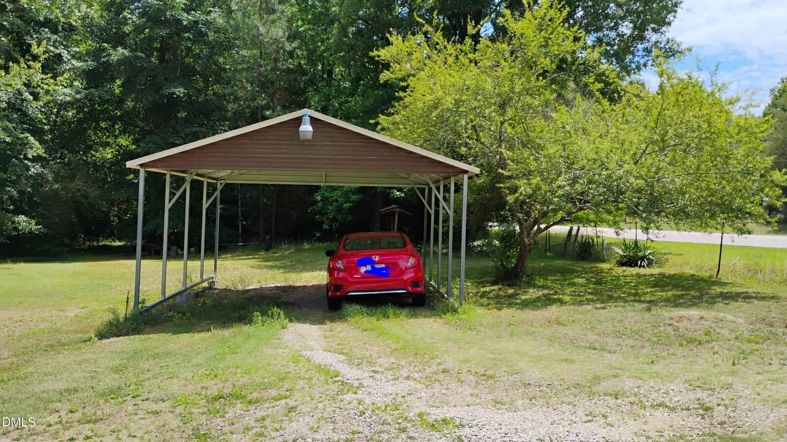 A red car is parked under a metal carport on a grassy lot with trees in the background and a gravel driveway leading up to the structure.