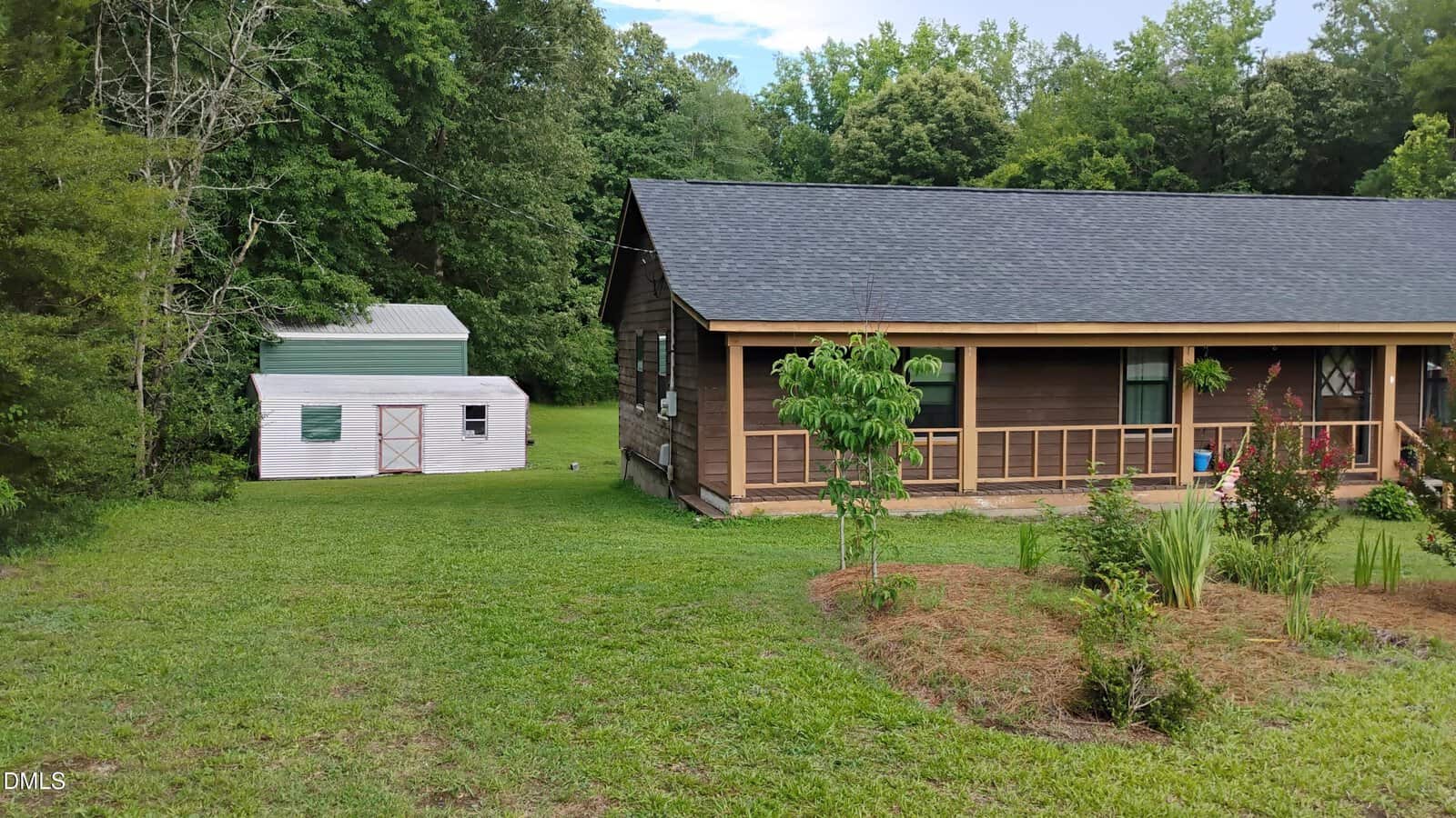 A wooden house with a covered porch stands on a grassy lawn. A small white shed is in the background, surrounded by green trees and shrubs.