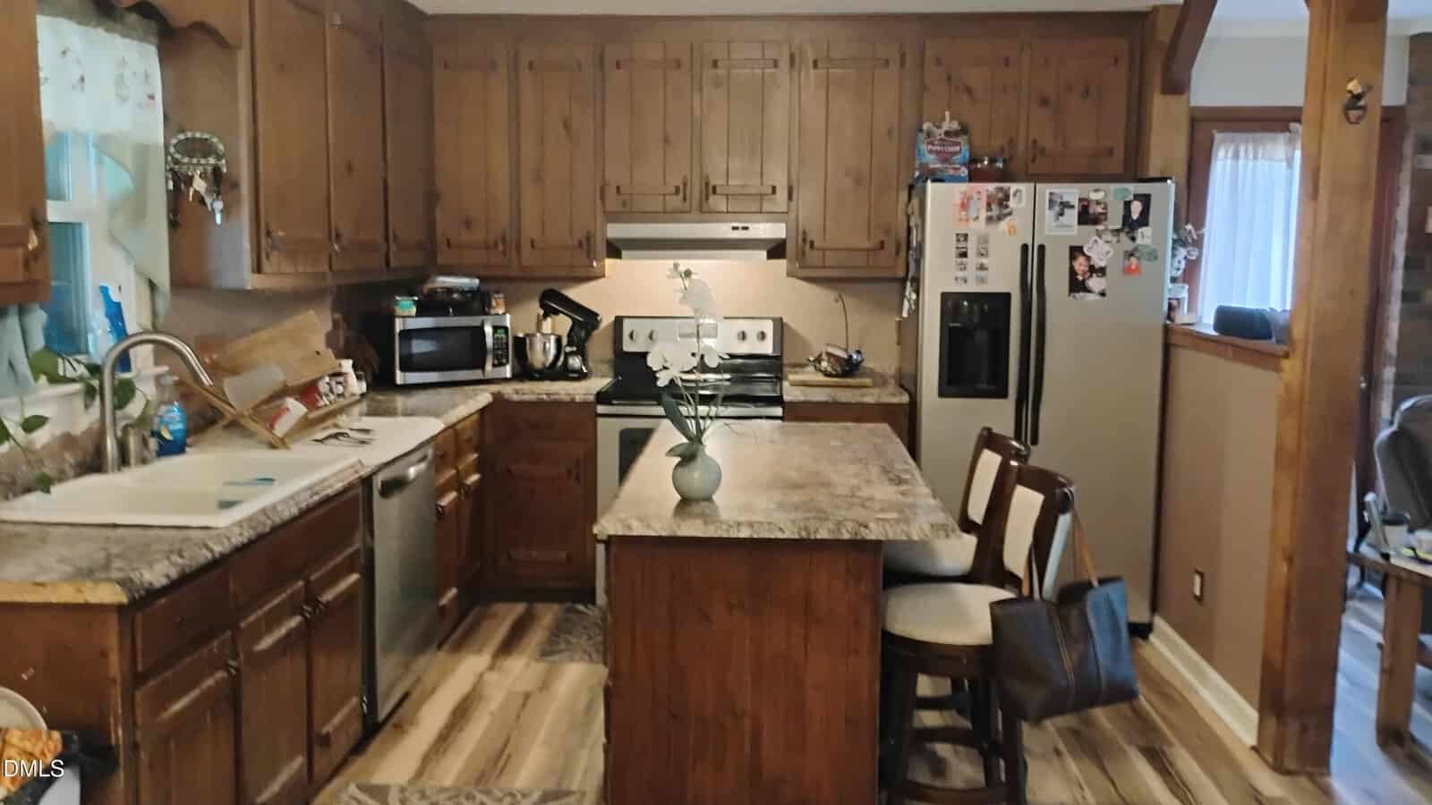 A cozy kitchen with wooden cabinets, a center island with chairs, stainless steel appliances, a vase with white flowers on the island, and various items on the counters and refrigerator.