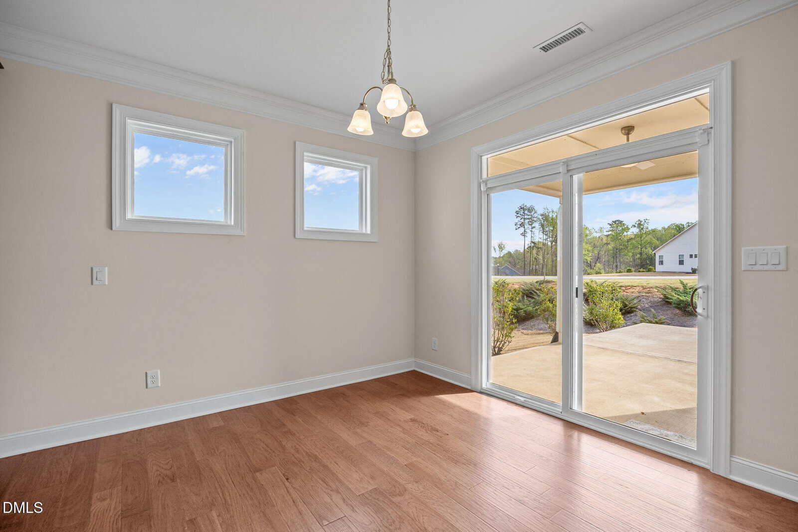 A bright, empty room with wood floors, two small windows, a hanging light fixture, and glass doors leading to a patio and green outdoor area.