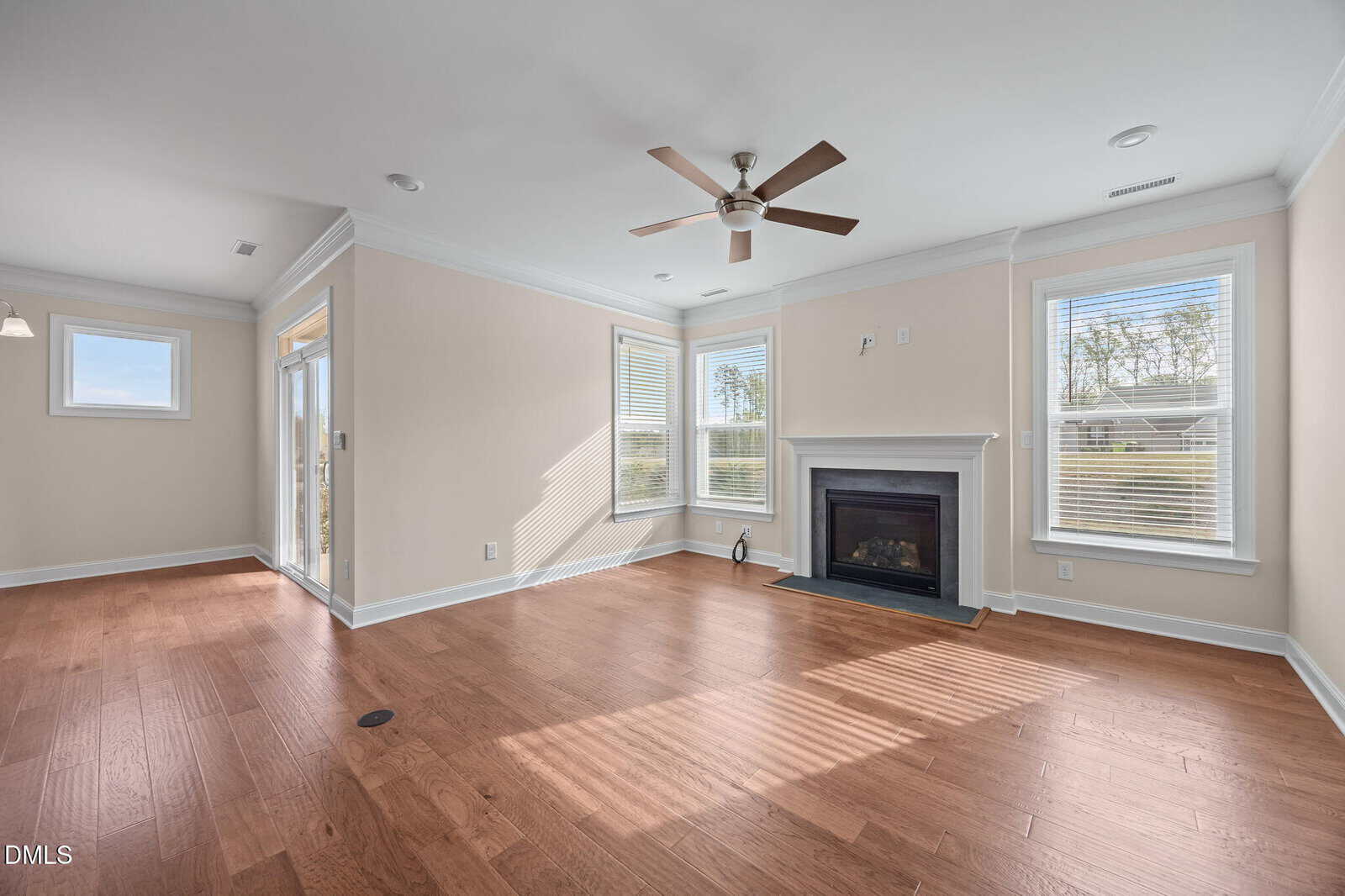 Bright living room with hardwood floors, beige walls, white trim, a ceiling fan, large windows, and a fireplace. An open doorway leads to a dining area with additional windows and natural light.