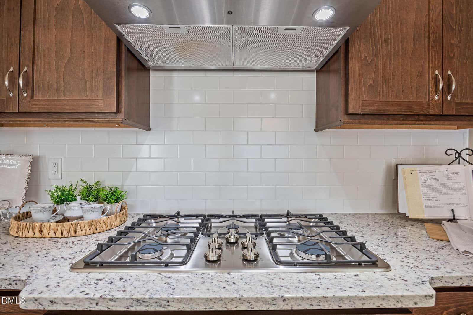 A kitchen stovetop with five burners sits below a stainless steel range hood, surrounded by wooden cabinets, white subway tile backsplash, and a granite countertop with cups, saucers, and an open recipe book.
