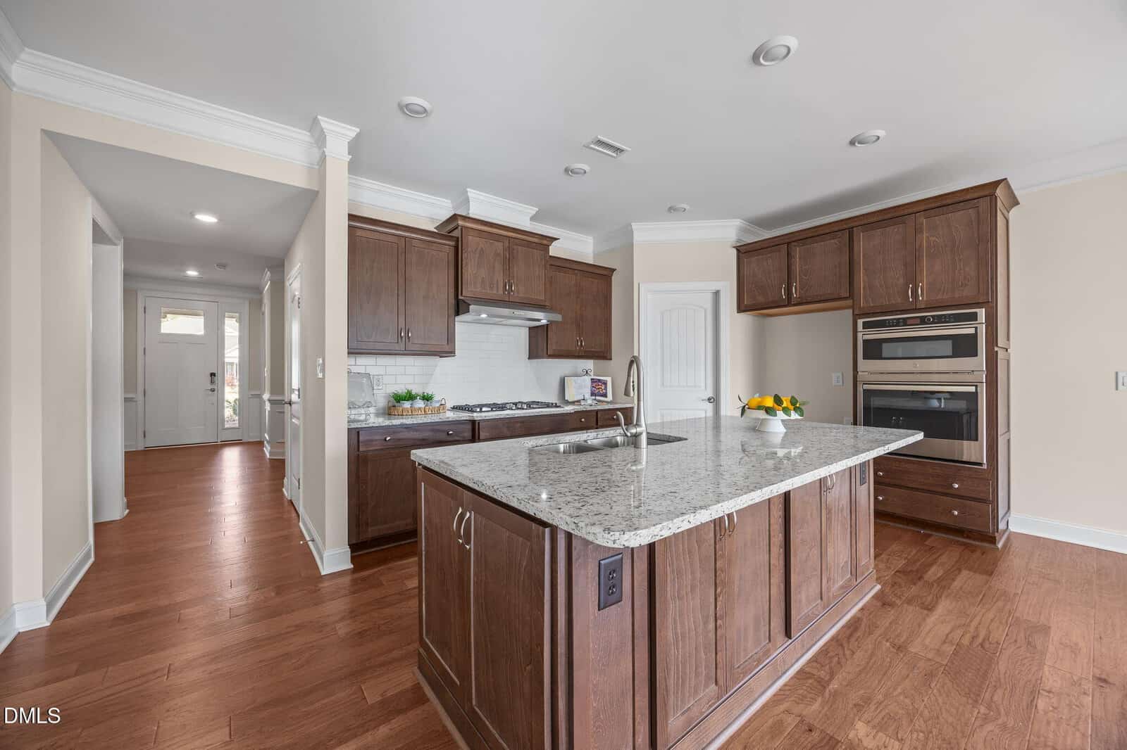 Modern kitchen with dark wood cabinets, granite island, built-in oven, gas stove, white subway tile backsplash, and hardwood floors. Bowl of lemons and decorative items are on the counters. Hallway and front door visible in background.