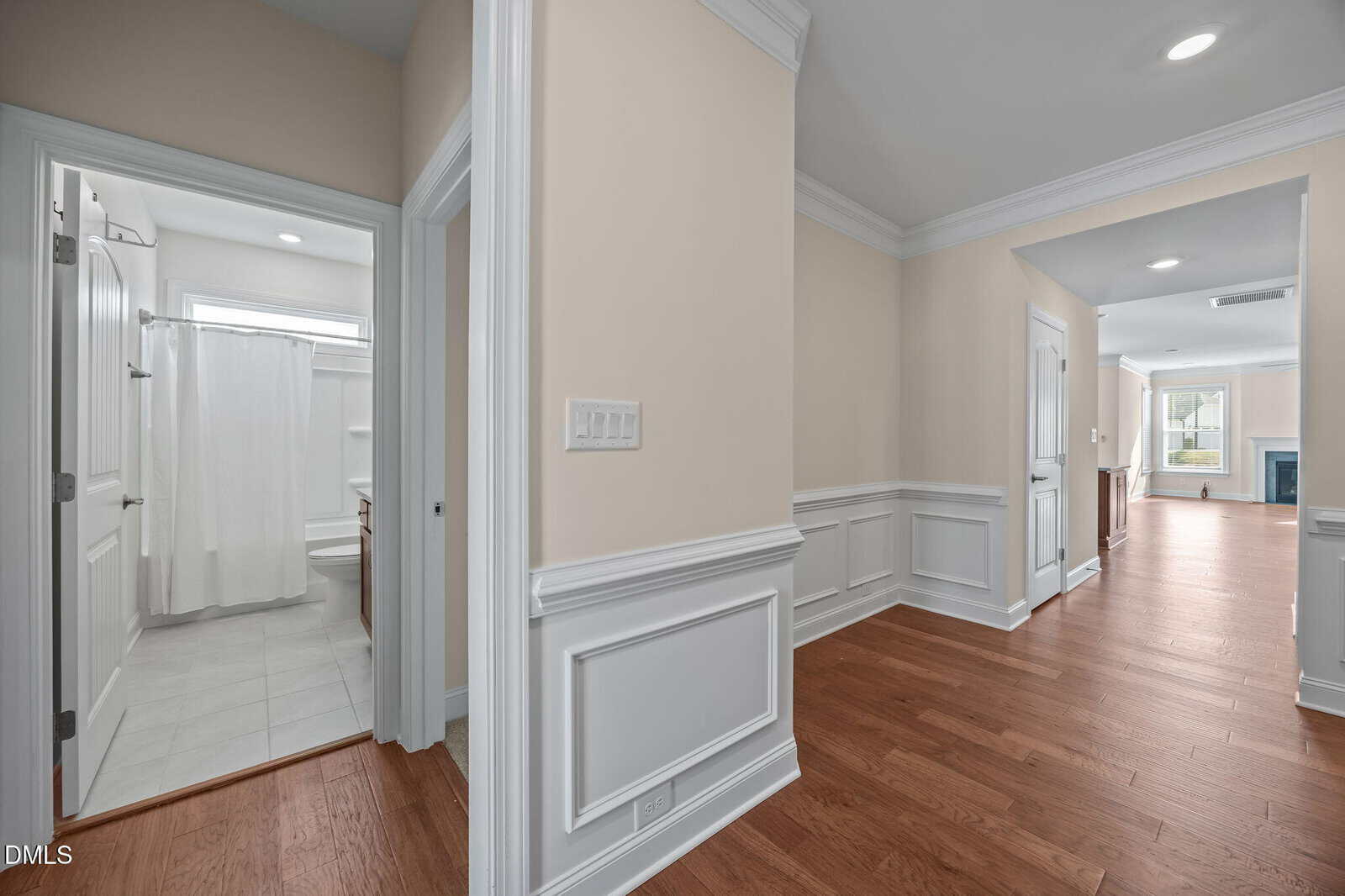 A hallway with wood floors and cream walls, white trim, and wainscoting. On the left is a bathroom with white fixtures and a shower curtain. The hallway leads to a bright living room with a fireplace.