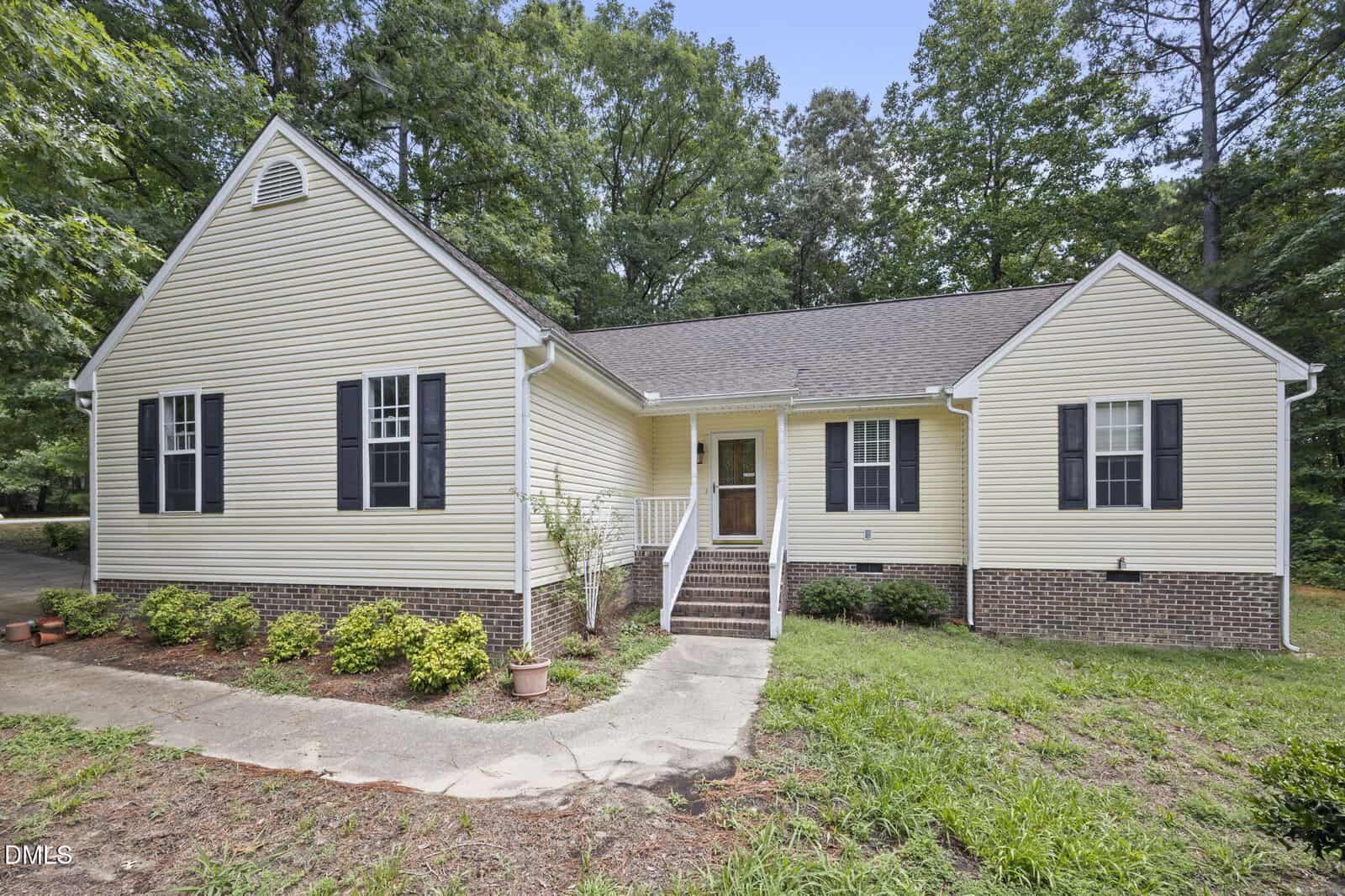 Charming single-story beige house at 235 Cherrylaurel Drive, Youngsville, featuring black shutters, a gable roof, brick foundation, front steps with white railings, and surrounded by lush shrubs, mature trees, and a grassy yard.