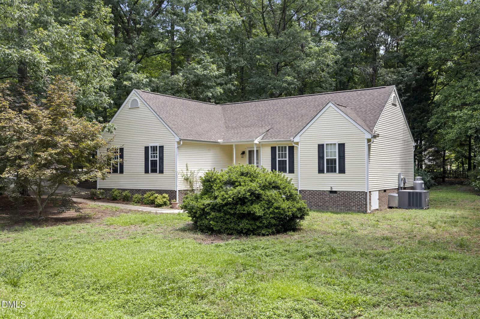 Single-story yellow house with white trim, dark shutters, and a gable roof, surrounded by trees and bushes, with a green lawn in front.