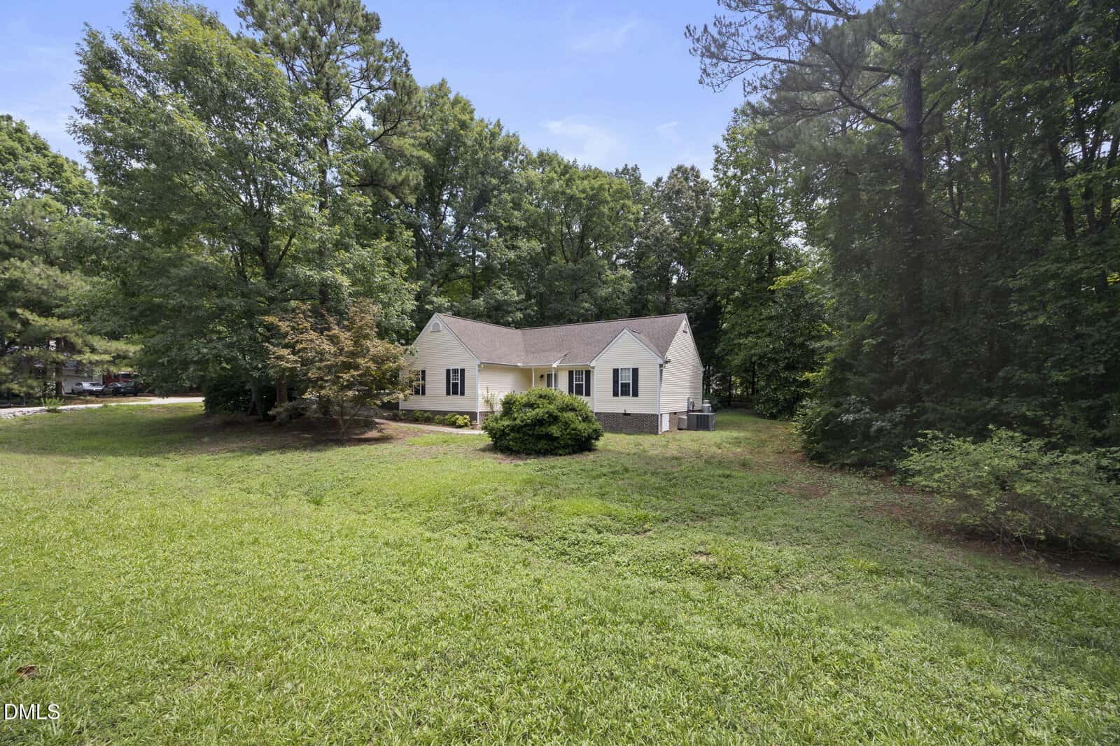 Single-story white house with a gray roof, surrounded by green grass and trees on a sunny day. The house sits in the center of a spacious, open yard bordered by woods.
