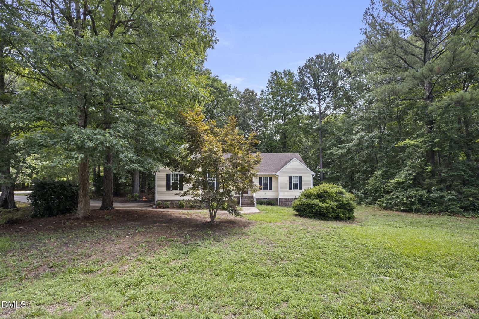 Single-story white house with a dark roof sits among tall trees, surrounded by green grass and shrubs under a clear blue sky. A small path leads to the front porch.