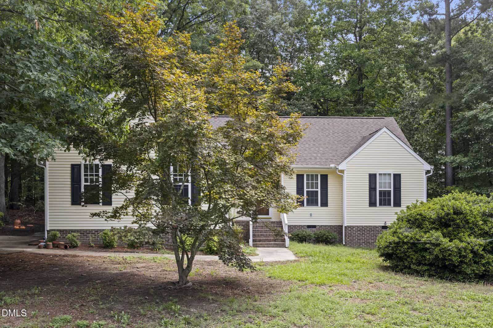 Single-story beige house with white trim, black shutters, and a gabled roof surrounded by trees, with a large leafy tree and bushes in the front yard. Steps lead to the front door.
