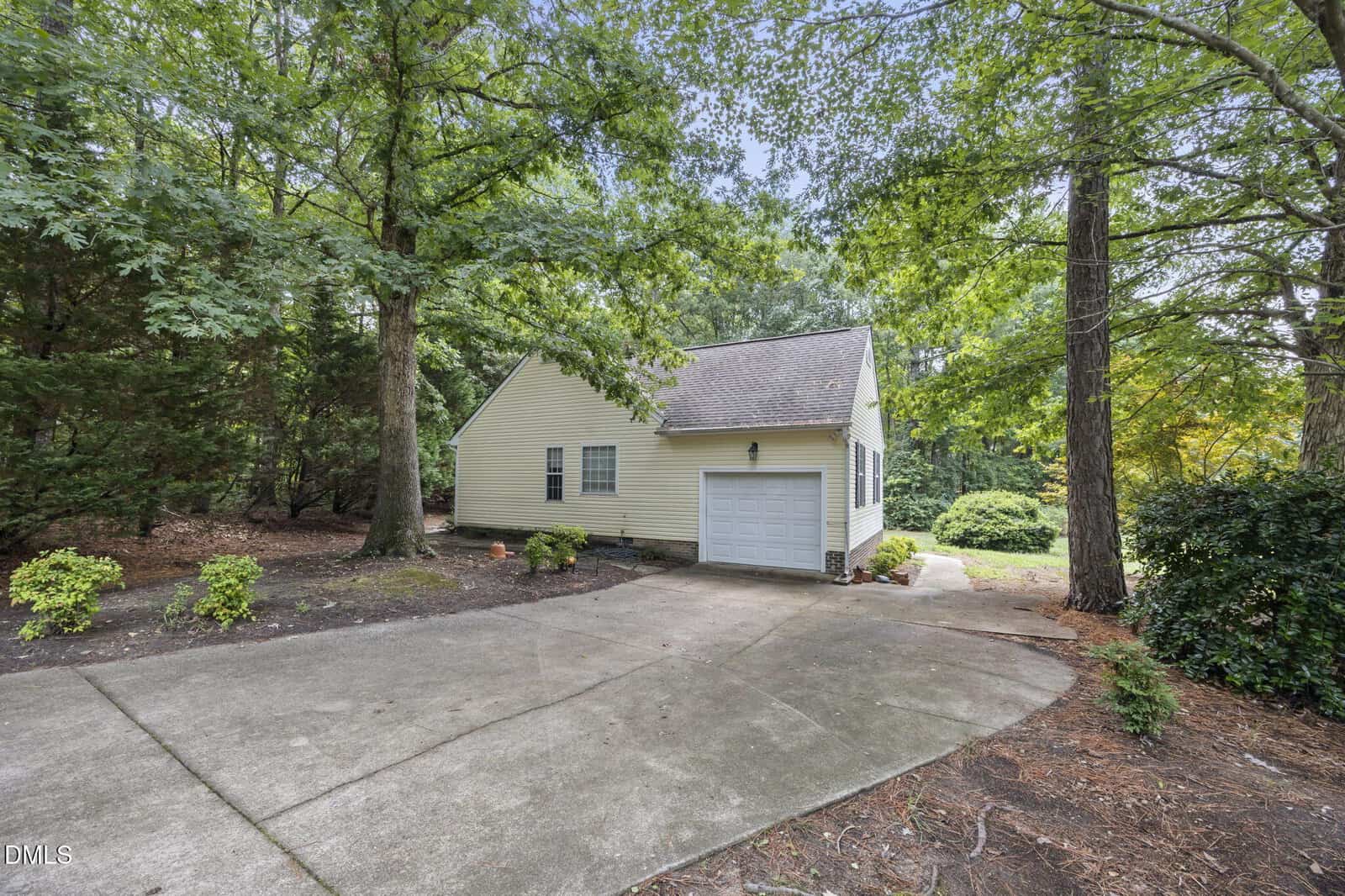 A small beige house with a single-car garage sits among tall trees, with a concrete driveway and minimal landscaping surrounding it.