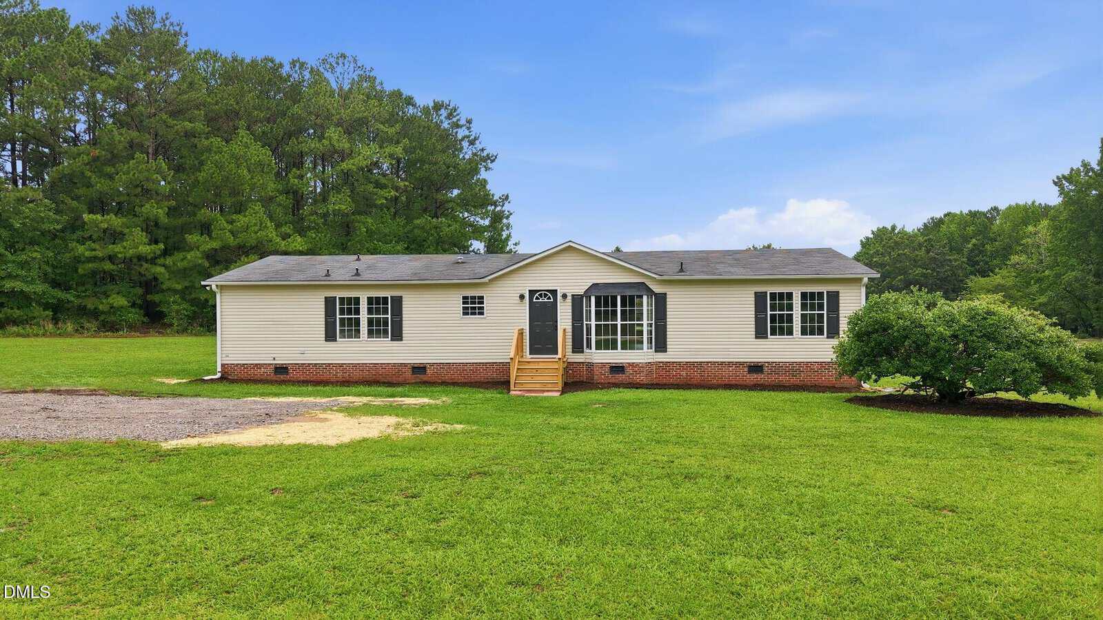 A single-story white house with a brick foundation, front steps, and large front windows sits on a green lawn with trees in the background under a partly cloudy sky.