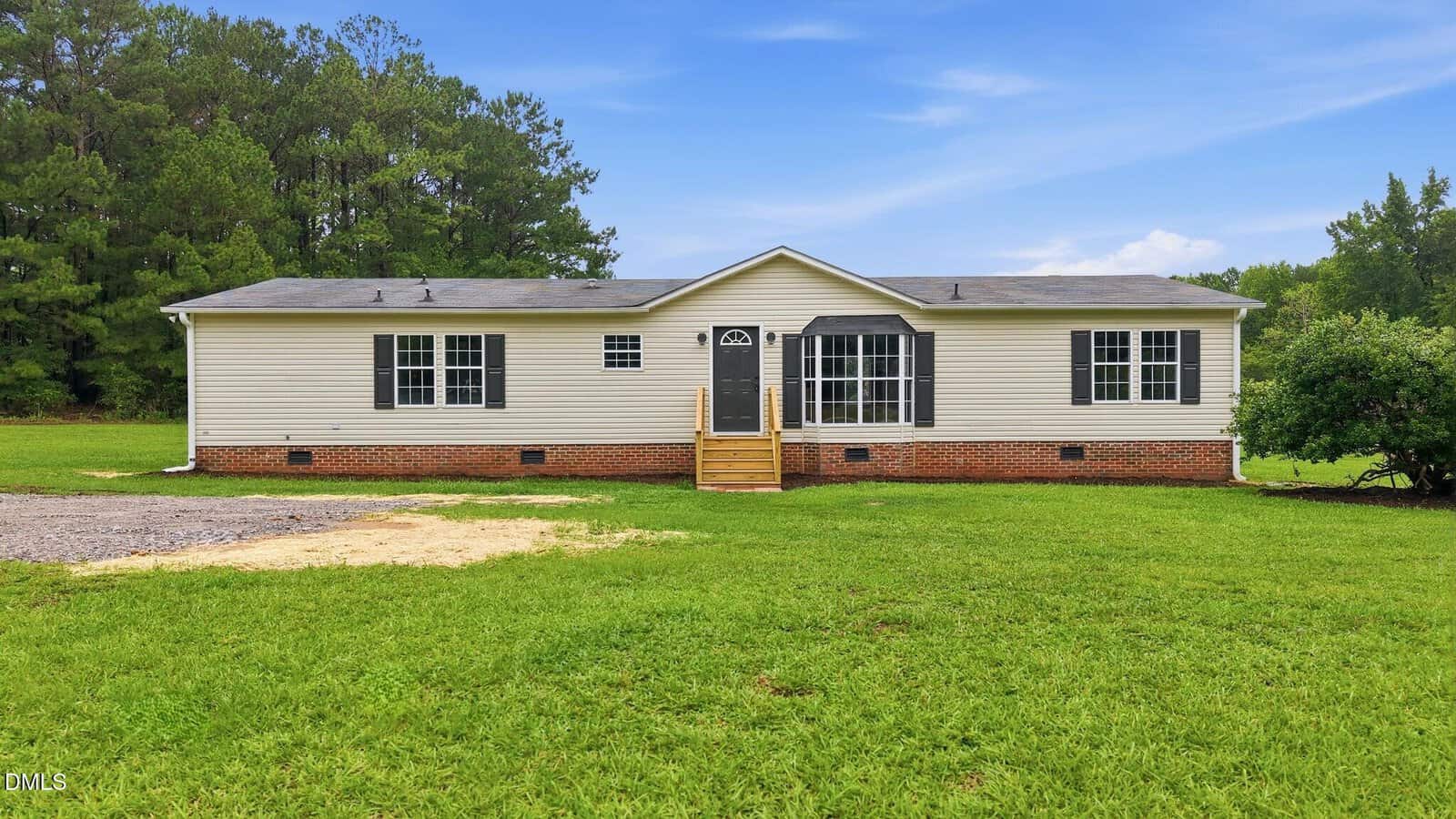 Single-story beige house with black shutters, a dark front door, small porch steps, and surrounded by green grass with trees in the background under a blue sky.