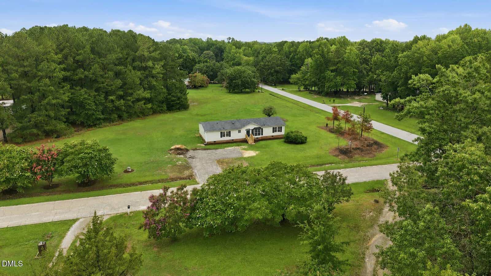 Aerial view of a single-story house with a gray roof, surrounded by spacious green lawns, trees, and roads in a quiet, wooded suburban neighborhood under a blue sky.