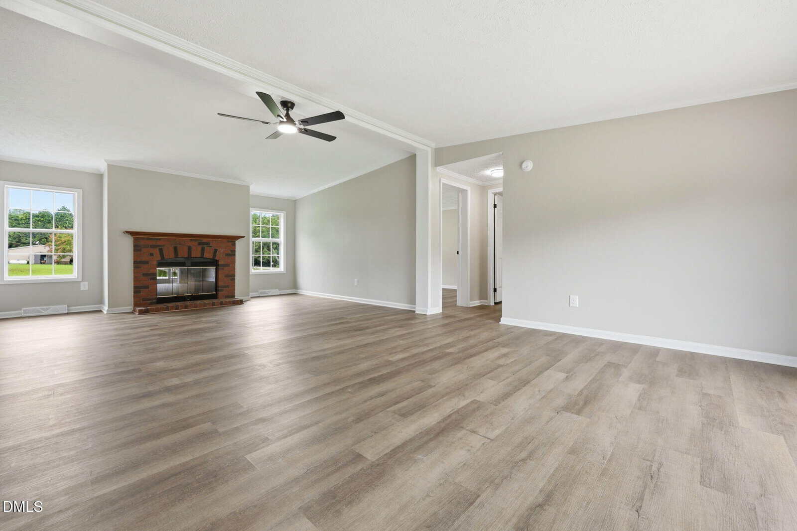 Spacious living room with light wood flooring, gray walls, a white ceiling, ceiling fan, large window, and a brick fireplace with a mantel. A hallway and open doorway are visible on the right.