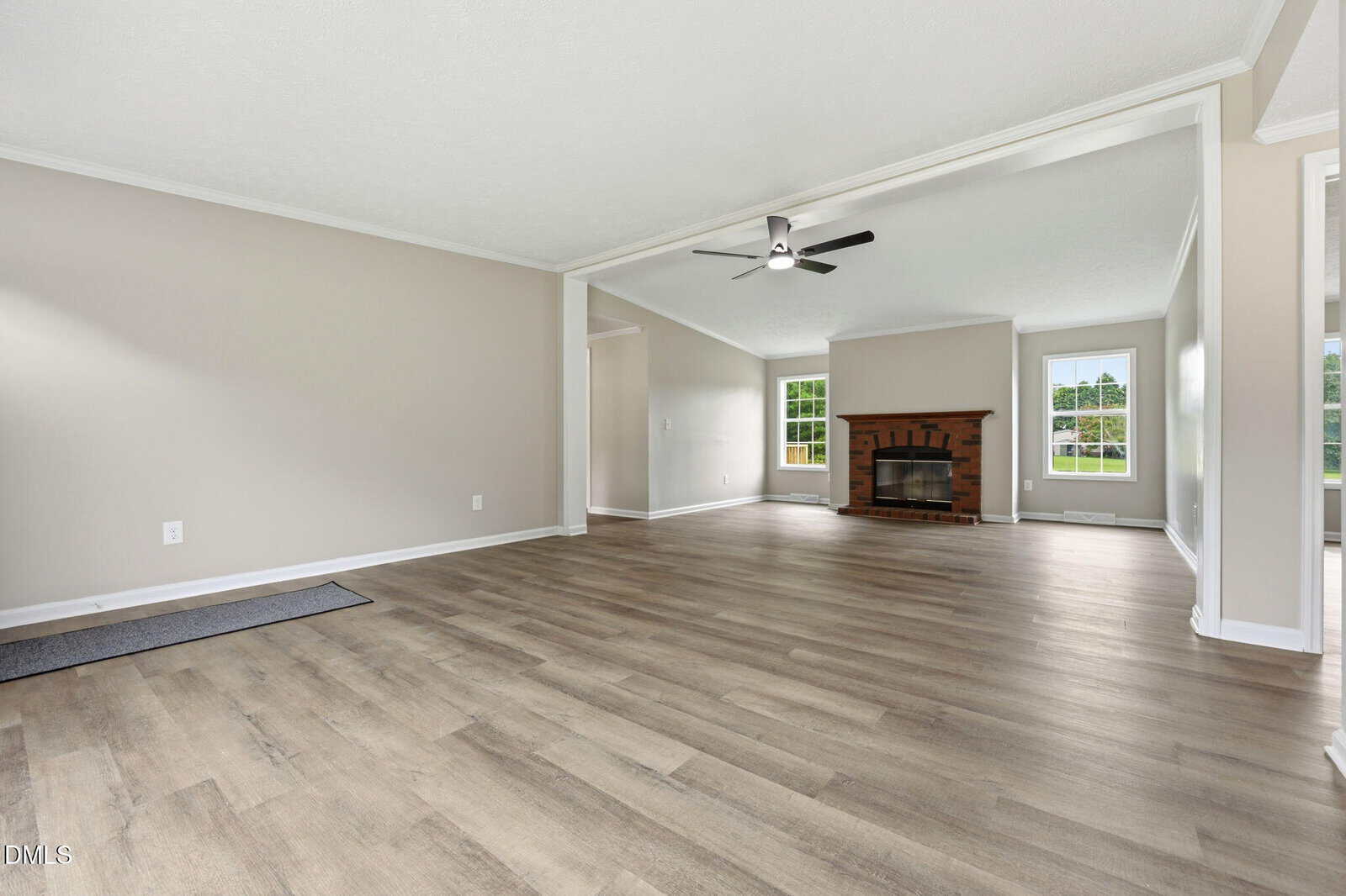 Spacious, empty living room with light wood flooring, beige walls, ceiling fan, large windows, and a brick fireplace in the background. Natural light fills the room.