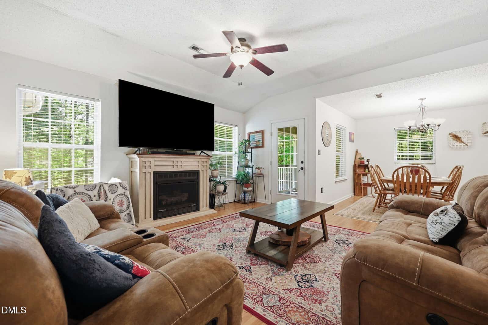 Cozy living room with two brown sofas, a coffee table, fireplace with TV above, and large windows. Dining area with wooden table and chairs is visible in the background. Ceiling fans and patterned rug complete the space.