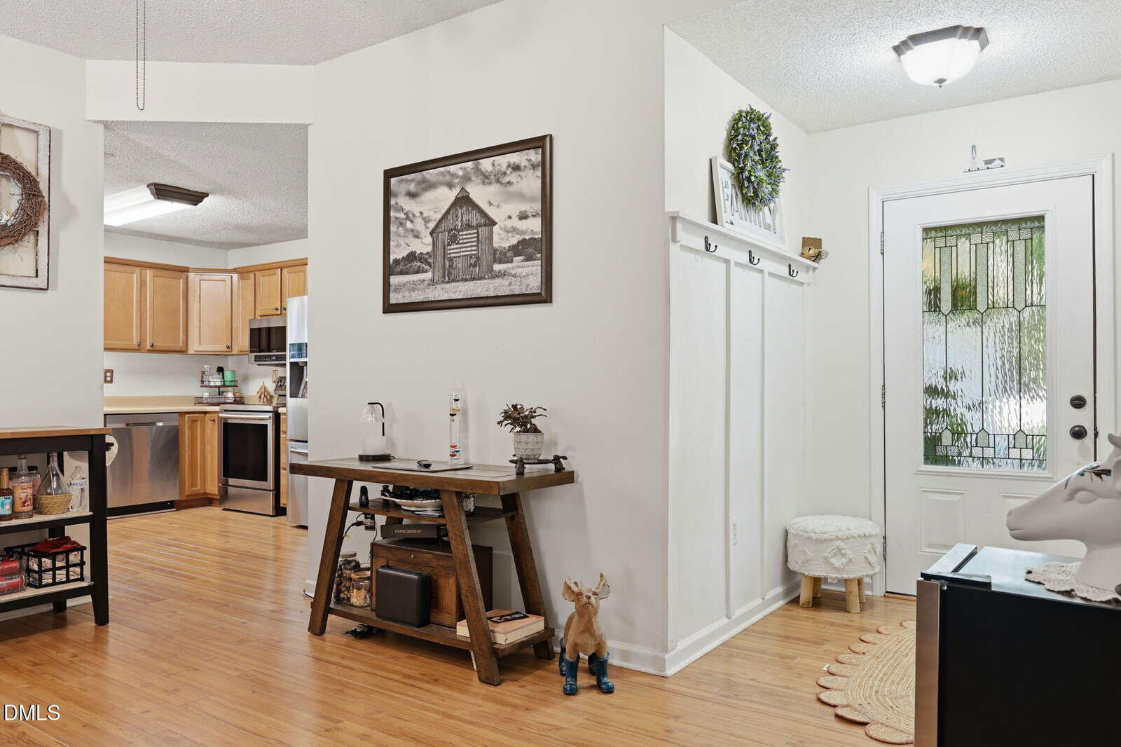 Entryway with a glass door, hooks, and a small bench. Adjacent room features wood floors, a rustic table, and a kitchen with light wood cabinets and stainless steel appliances in the background.