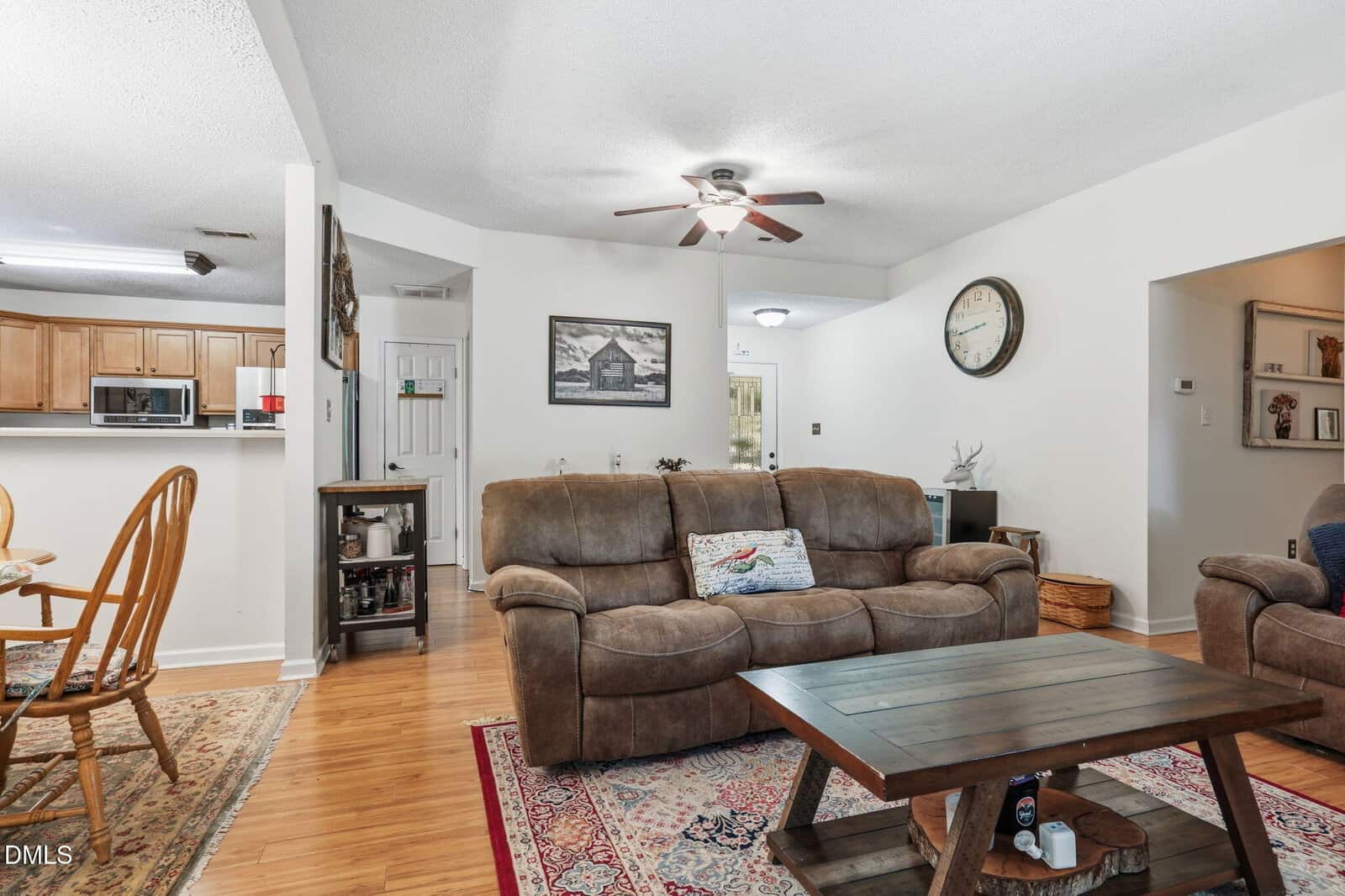 Cozy living room with brown sofa, wooden coffee table, wall clock, and kitchen in the background. Area rugs cover hardwood floors. Ceiling fan with light is on. A framed picture hangs on the wall above the sofa.
